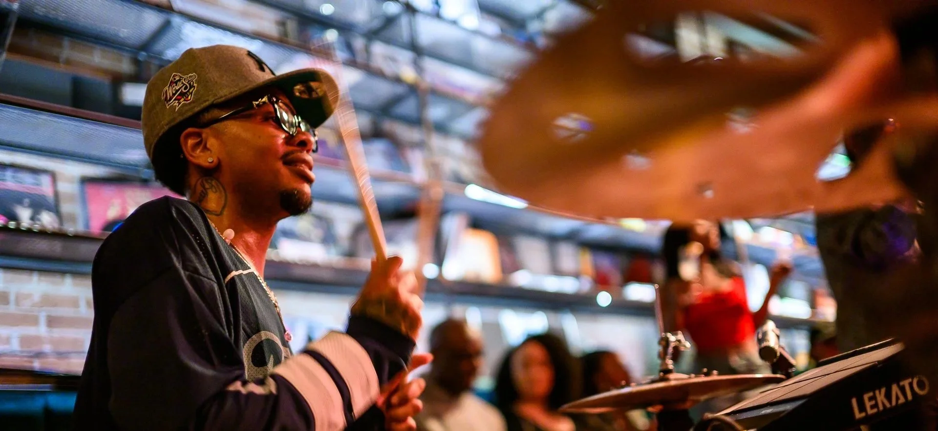 A man wearing sunglasses, a gray cap with a logo, and a black jacket playing drums in a lively indoor space with a brick wall and posters in the background.