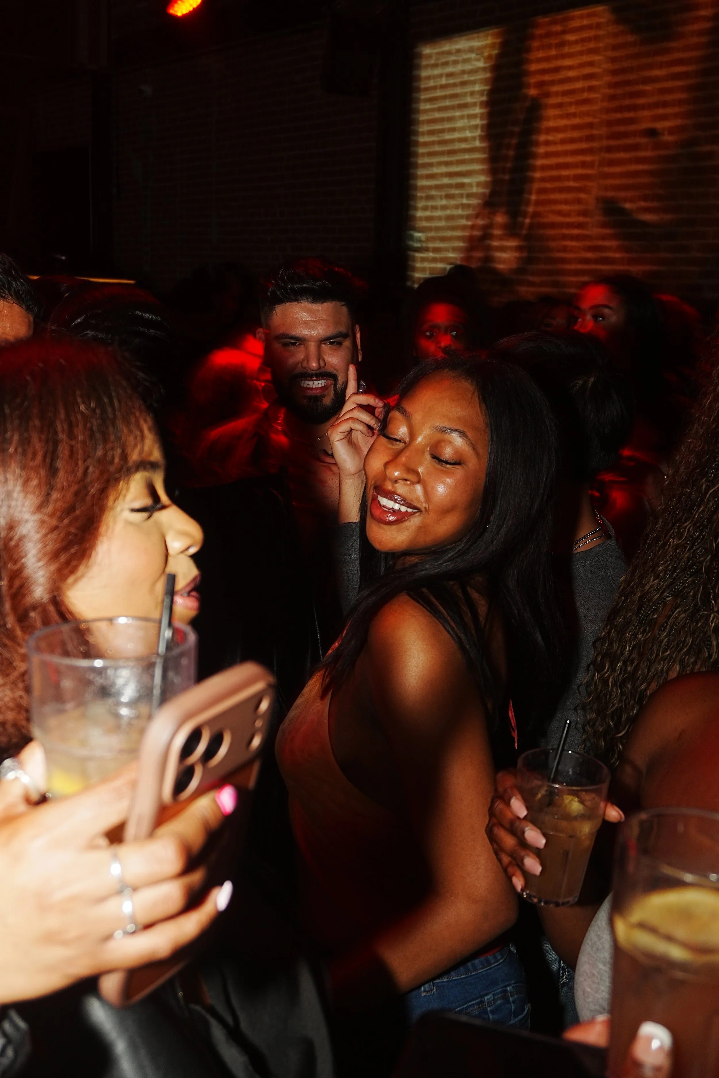 Group of people enjoying a party with drinks, featuring a woman in the center smiling with eyes closed holding a drink, and a man in the background smiling with a beard, in a dimly lit venue with exposed brick walls.