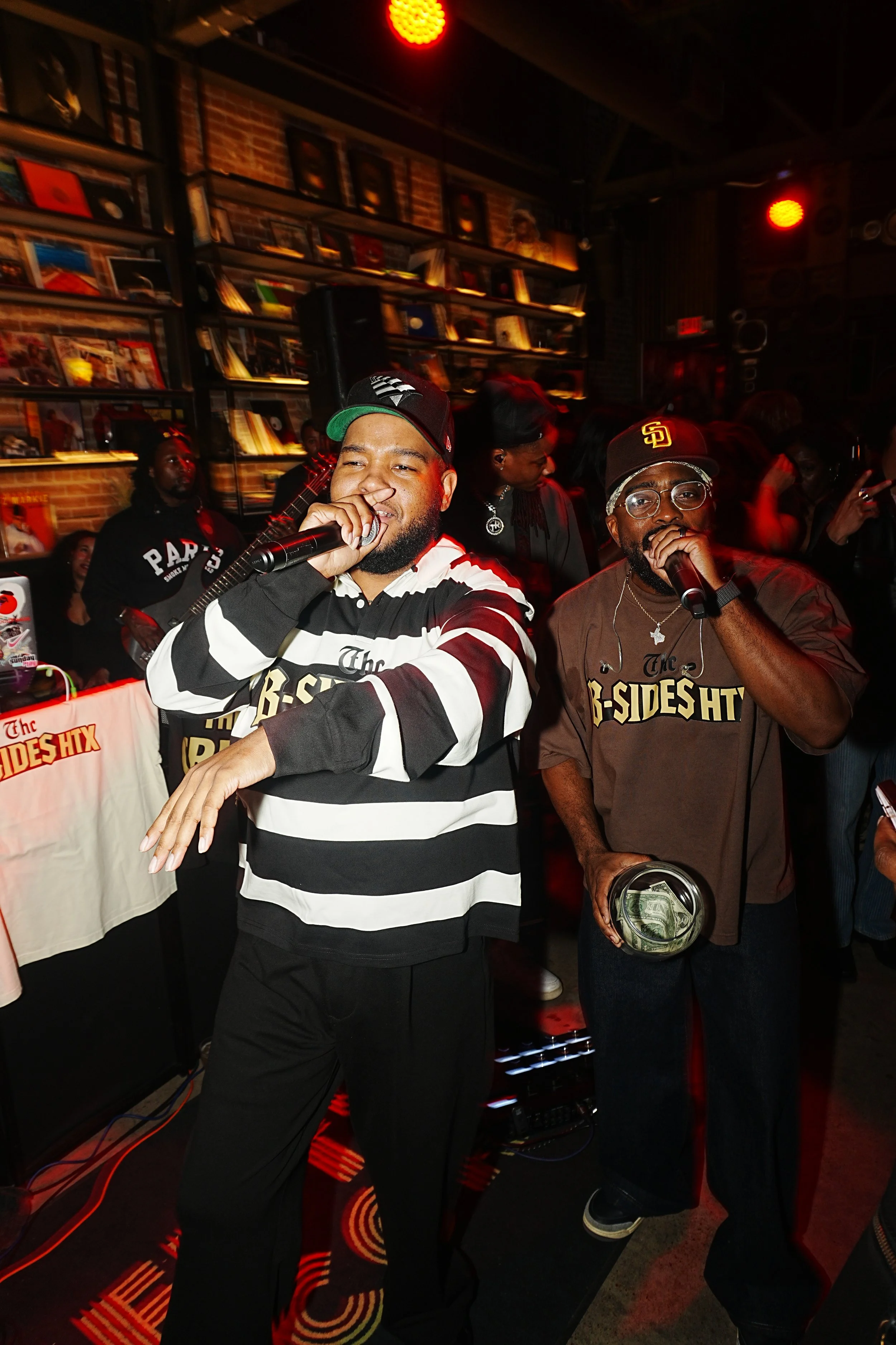 Two men singing or rapping into microphones at a lively indoor party or event, with a crowd and bookshelf in the background.