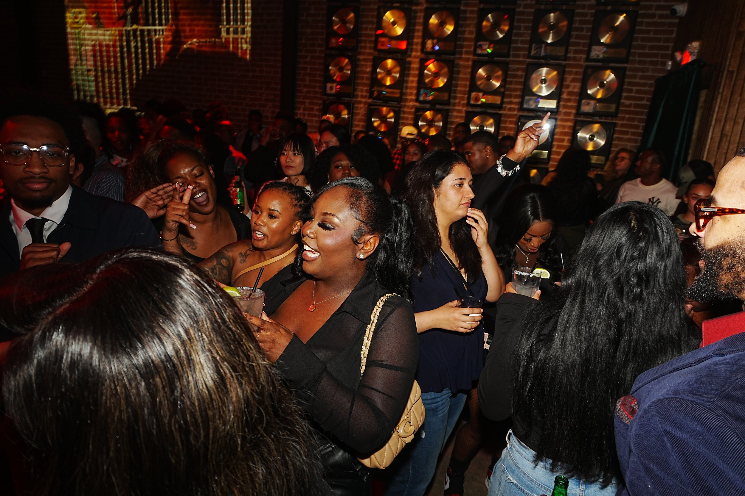 People dancing and socializing at a lively indoor event, with gold records on the brick wall in the background.