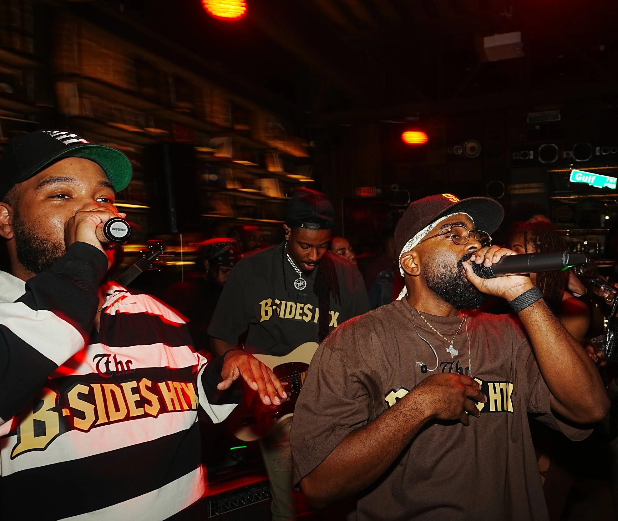 Three men performing at a music event, with one singing into a microphone, one playing guitar, and another holding a microphone near his mouth.