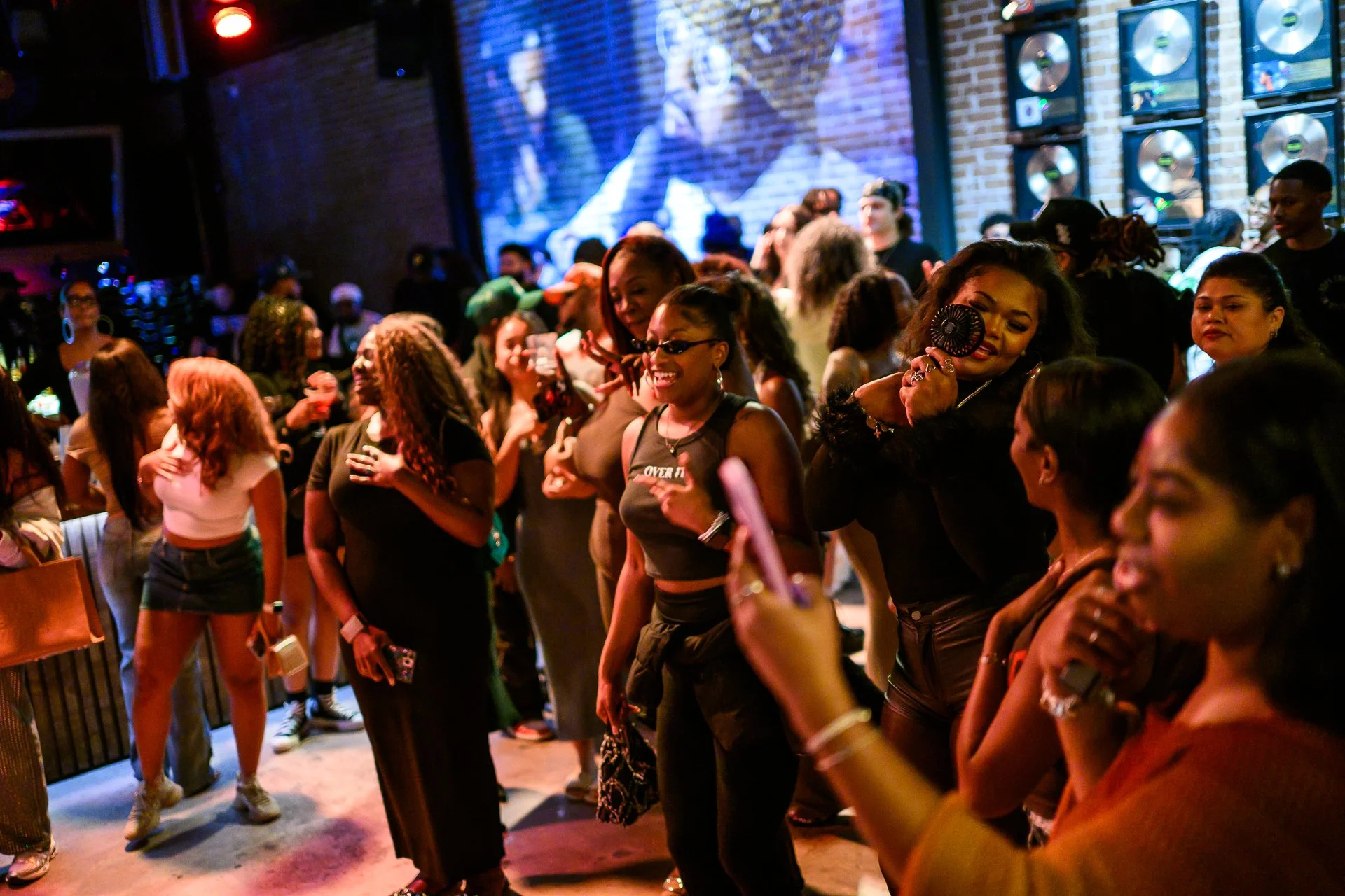 People dancing and socializing at a lively indoor concert or nightclub with a large screen displaying a video behind them.