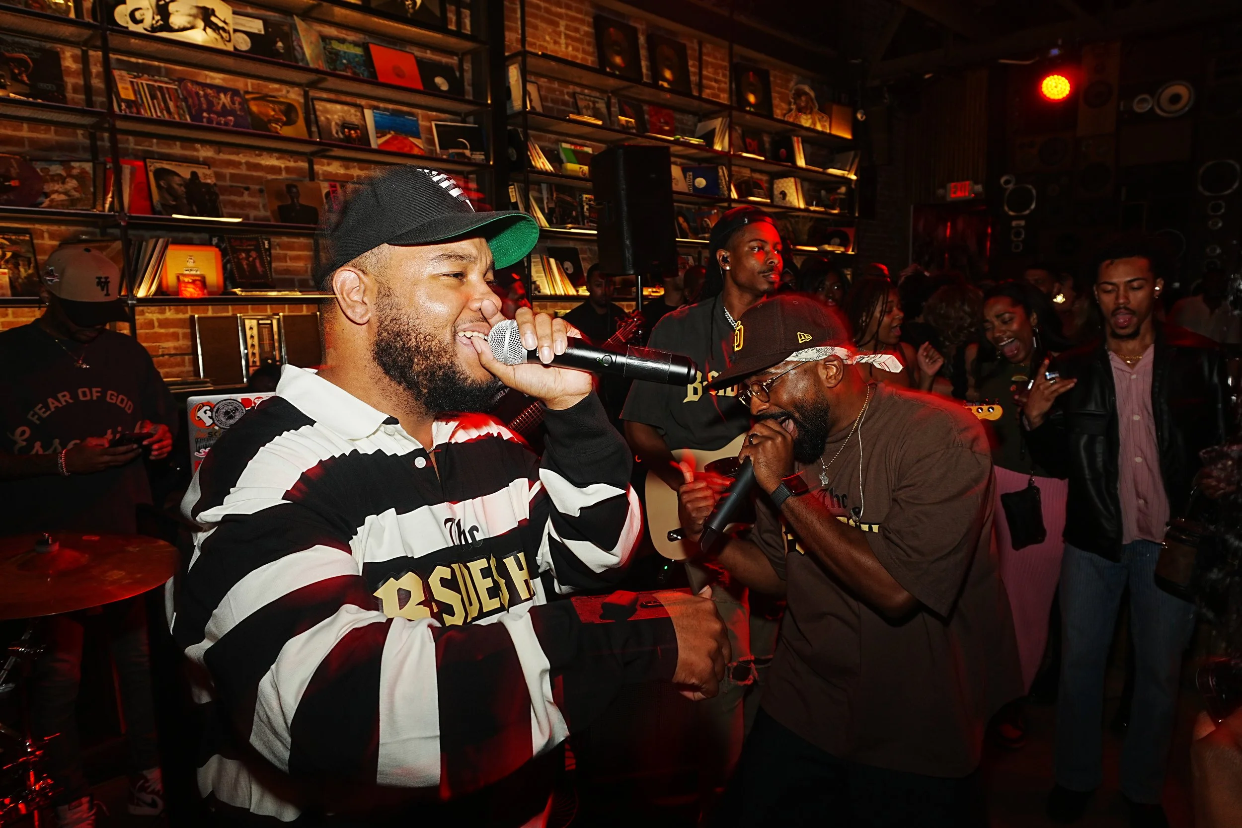 Two men singing into microphones at a lively indoor event with a crowd, brick wall decorated with shelves of vinyl records in the background.
