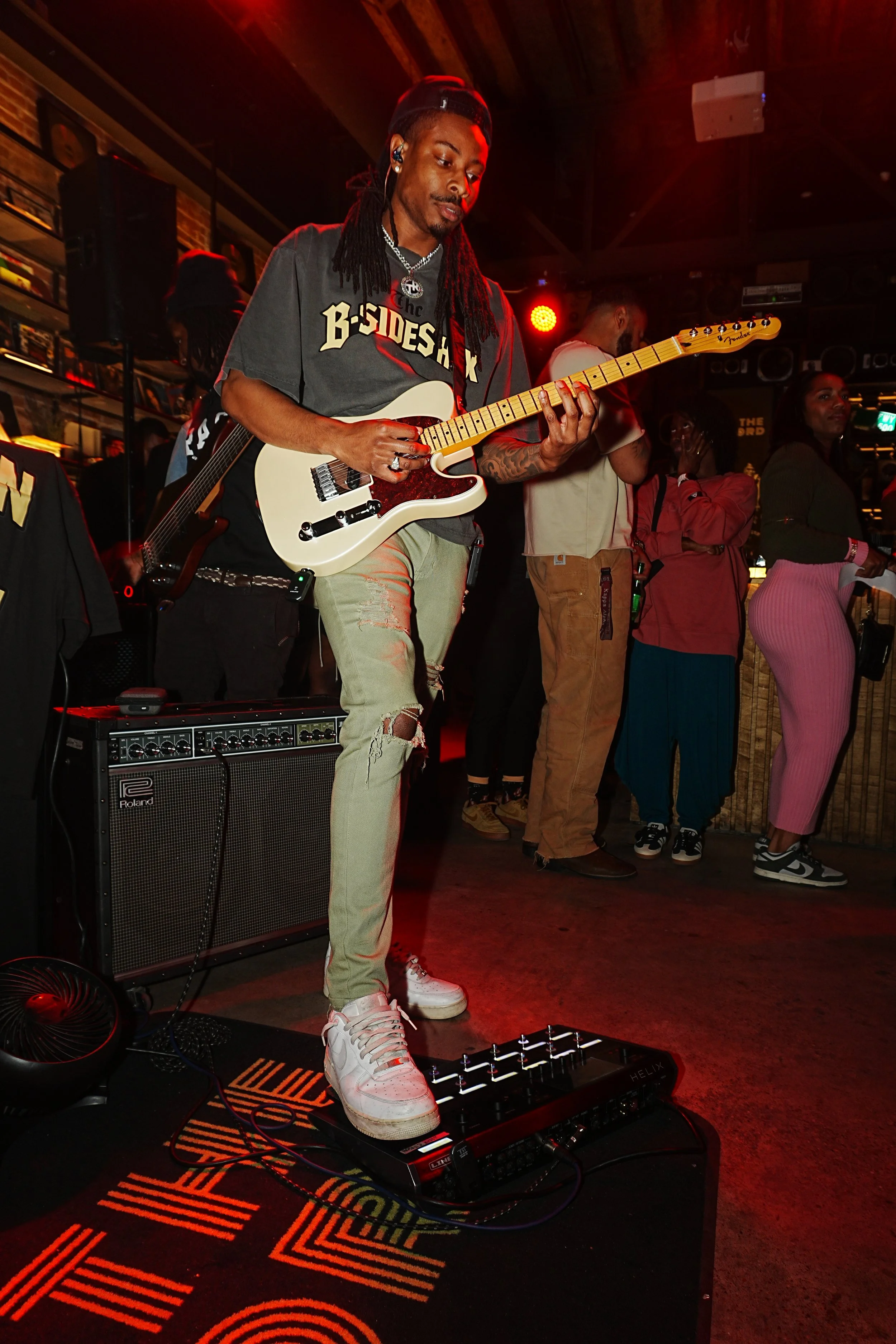 A musician with long dreadlocks playing a white electric guitar on stage in a bar or club, with a small amplifier at his feet and a group of people watching in the background.