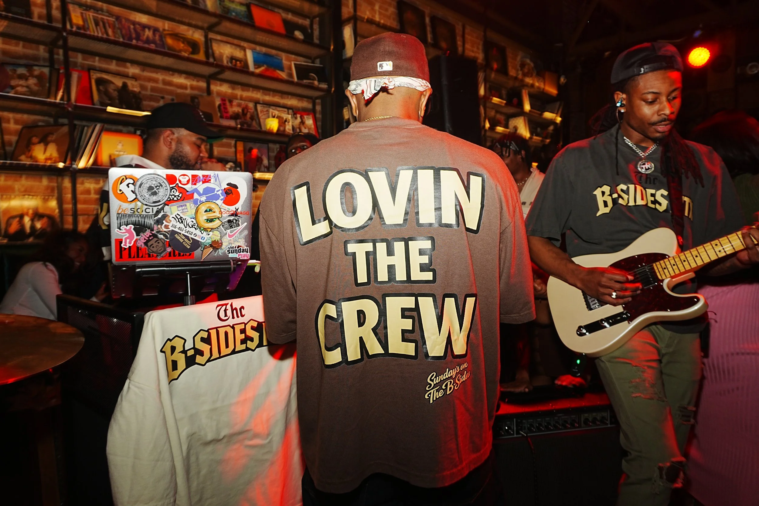 A man wearing a t-shirt with the words 'LOVIN THE CREW' seen from behind, standing next to a man playing an electric guitar at a lively indoor event, with shelves of books and framed photos on the wall in the background.