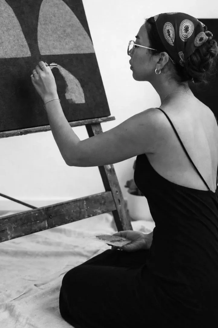 A woman painting on a canvas, sitting cross-legged on the floor, wearing glasses, a bandana, and a black dress.