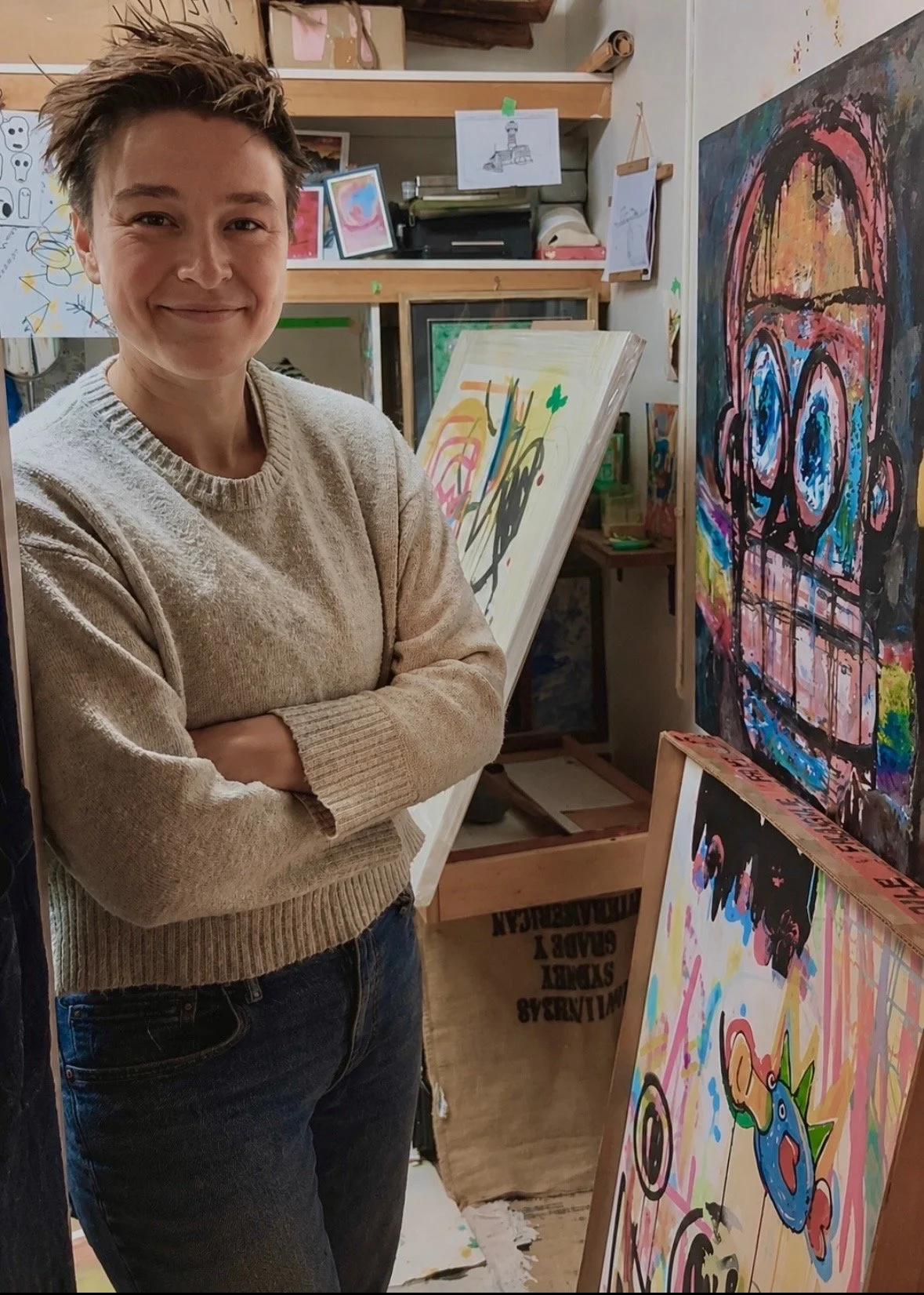 A person standing in an art studio with arms crossed, smiling, surrounded by colorful abstract paintings and artwork on shelves and walls.