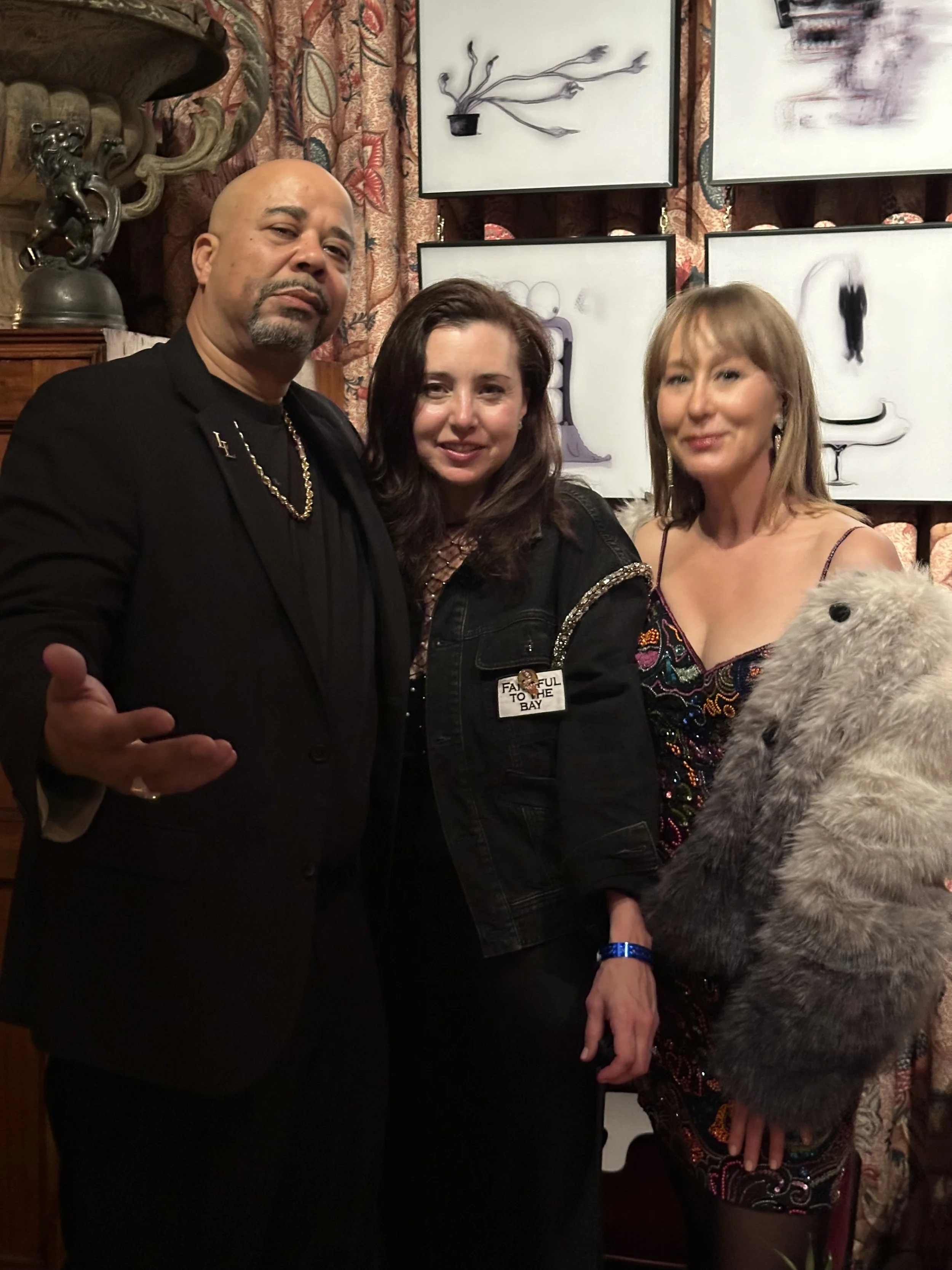 Three people standing together indoors with artwork on the wall behind them. The man on the left is wearing a black suit with a chain necklace, the woman in the middle has dark hair and is wearing a black jacket with a patch, and the woman on the rig