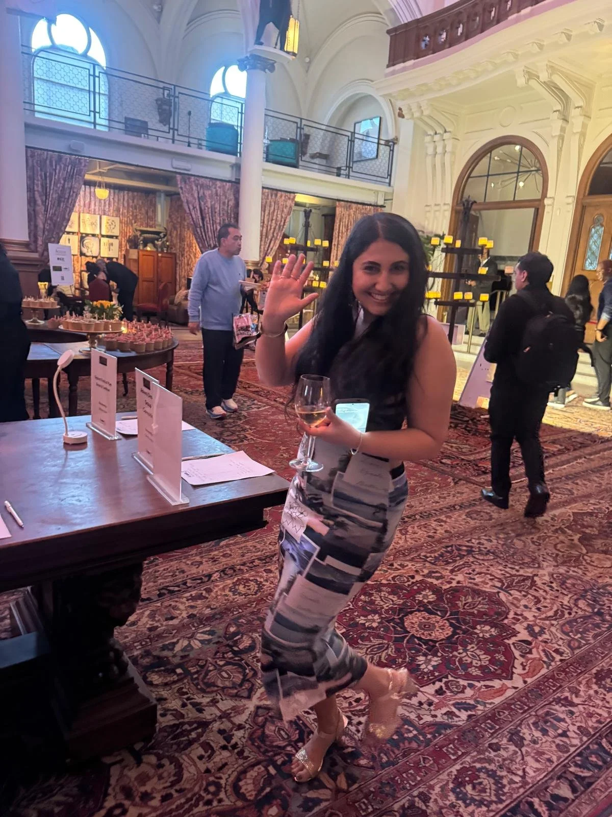 A woman with long dark hair smiling and waving, holding a glass of wine and a smartphone, standing in an elegant, historic hotel lobby with high ceilings, large windows, and ornate decor. Other people are seen in the background, and tables with small