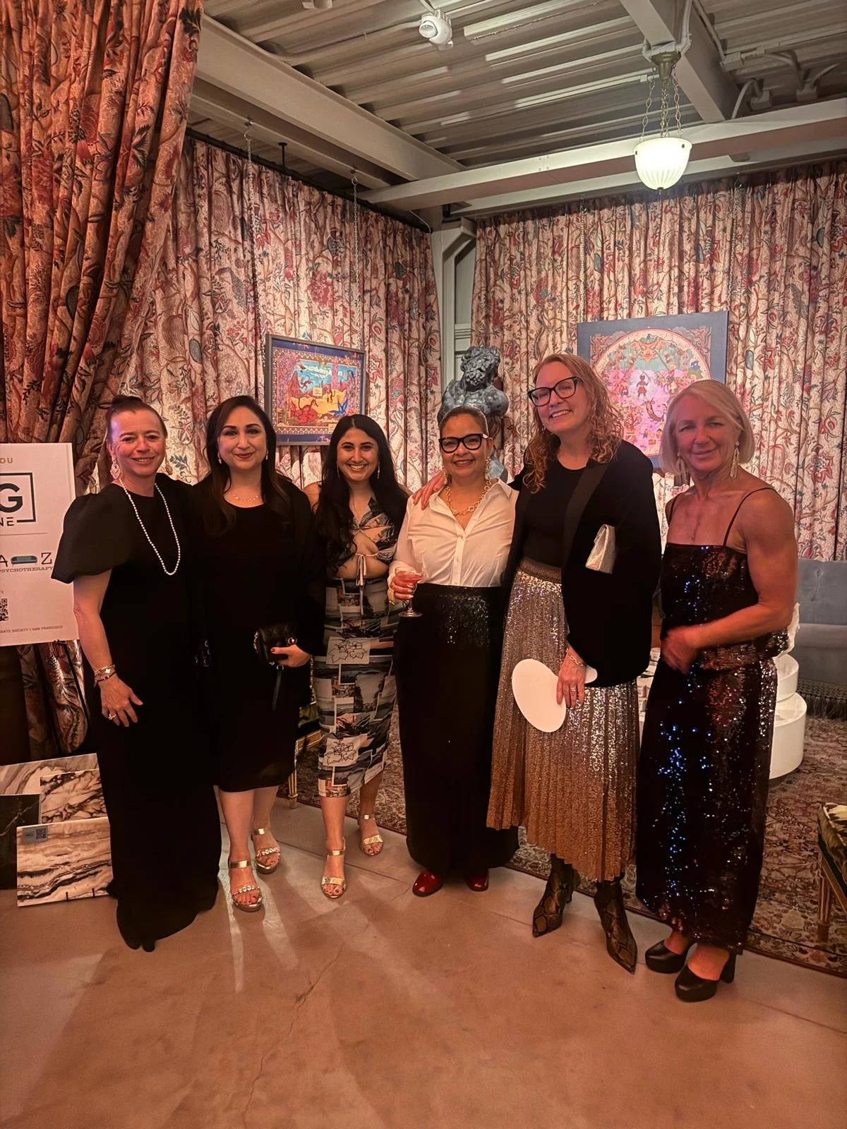 Six women in formal attire posing together in a decorated indoor space with patterned curtains and artwork in the background.
