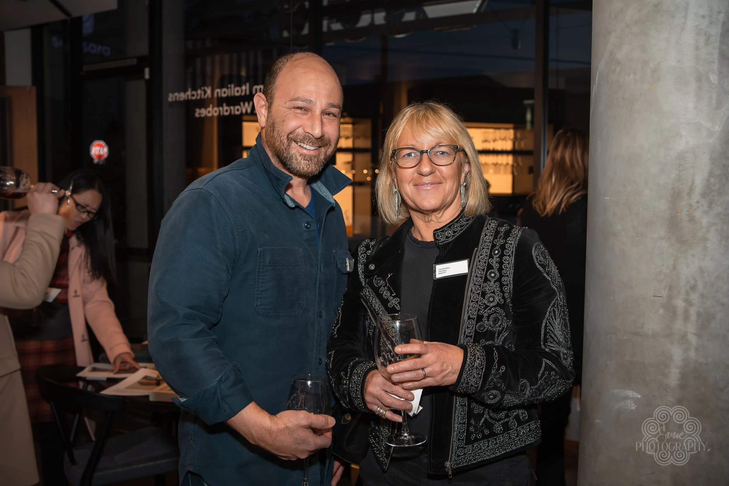 A man and a woman holding wine glasses at a social event, smiling and standing close together. The woman wears a black jacket with intricate embroidery, glasses, and earrings. The man has a beard and wears a dark blue shirt. In the background, there 