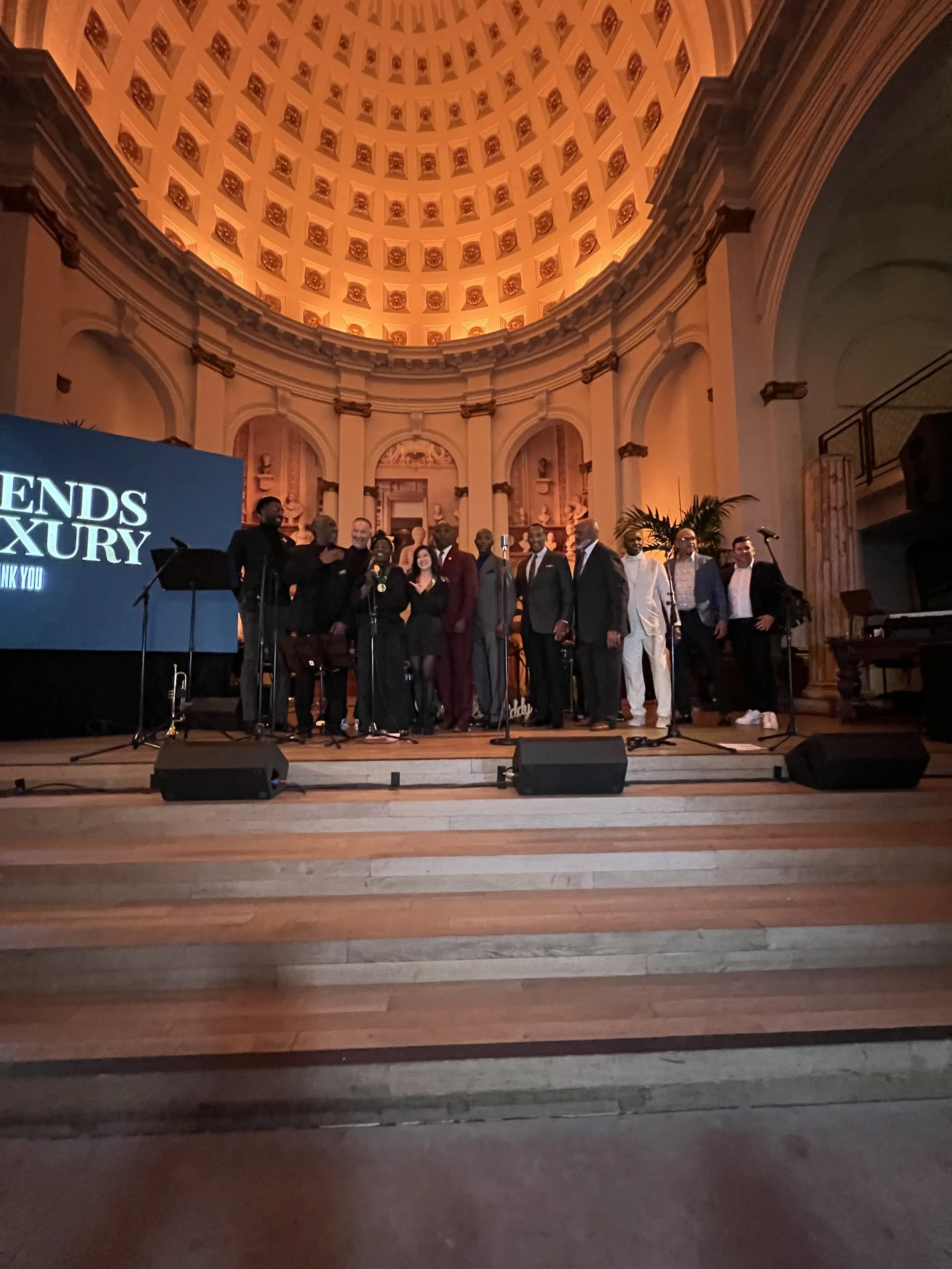 A group of people dressed in formal attire standing on a stage in a grand hall with a domed ceiling, microphones, and large screen with the words "ENDS LUXURY" in the background.