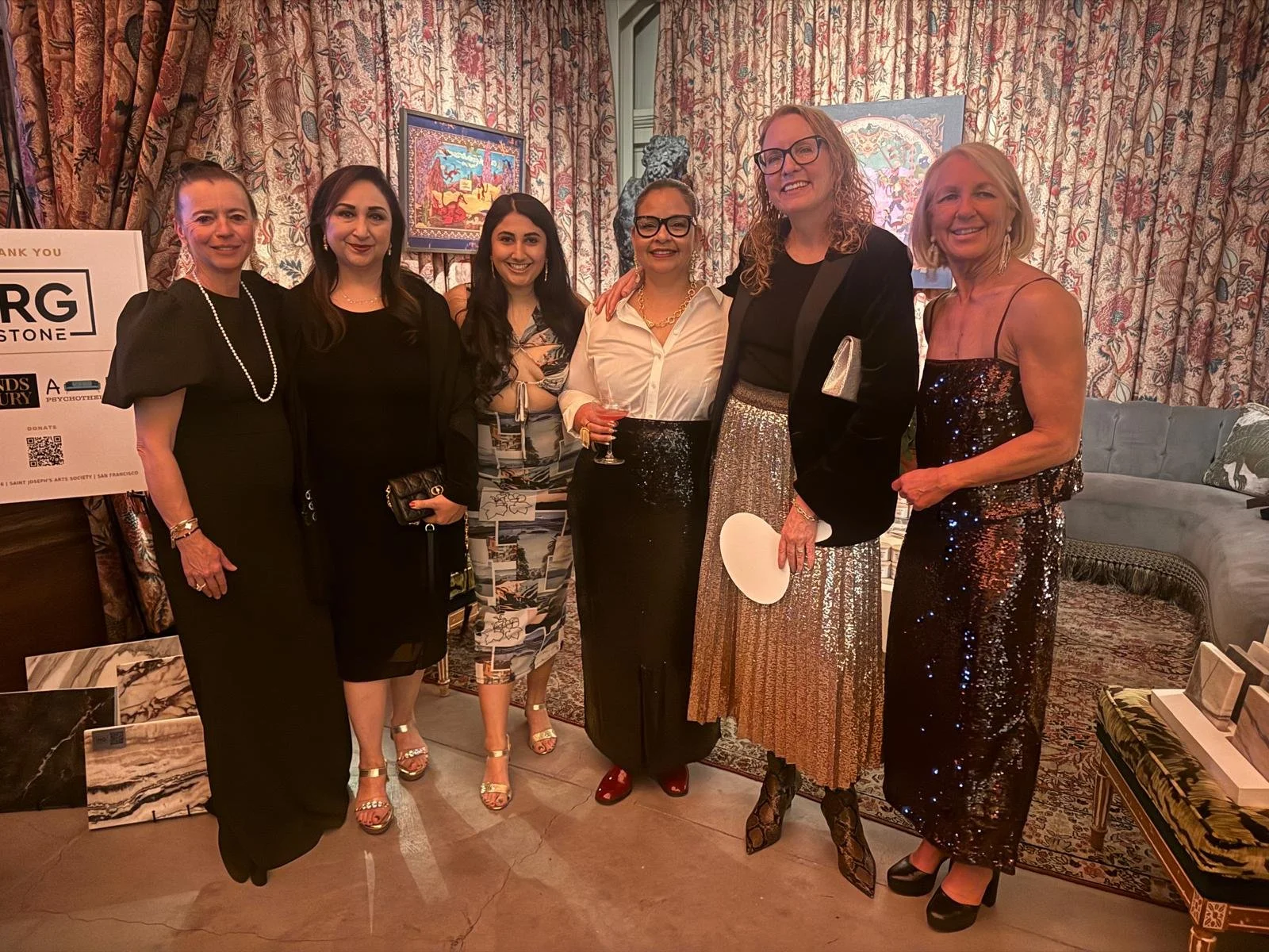 Group of seven women standing together indoors, dressed in formal and semi-formal attire, smiling at the camera.