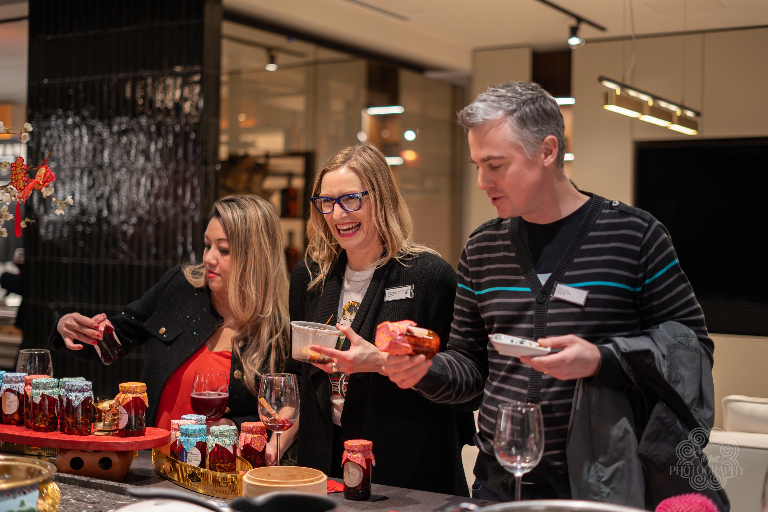 Three people smile and look at various jars and food items on a table at a market or event, with glasses of wine present.