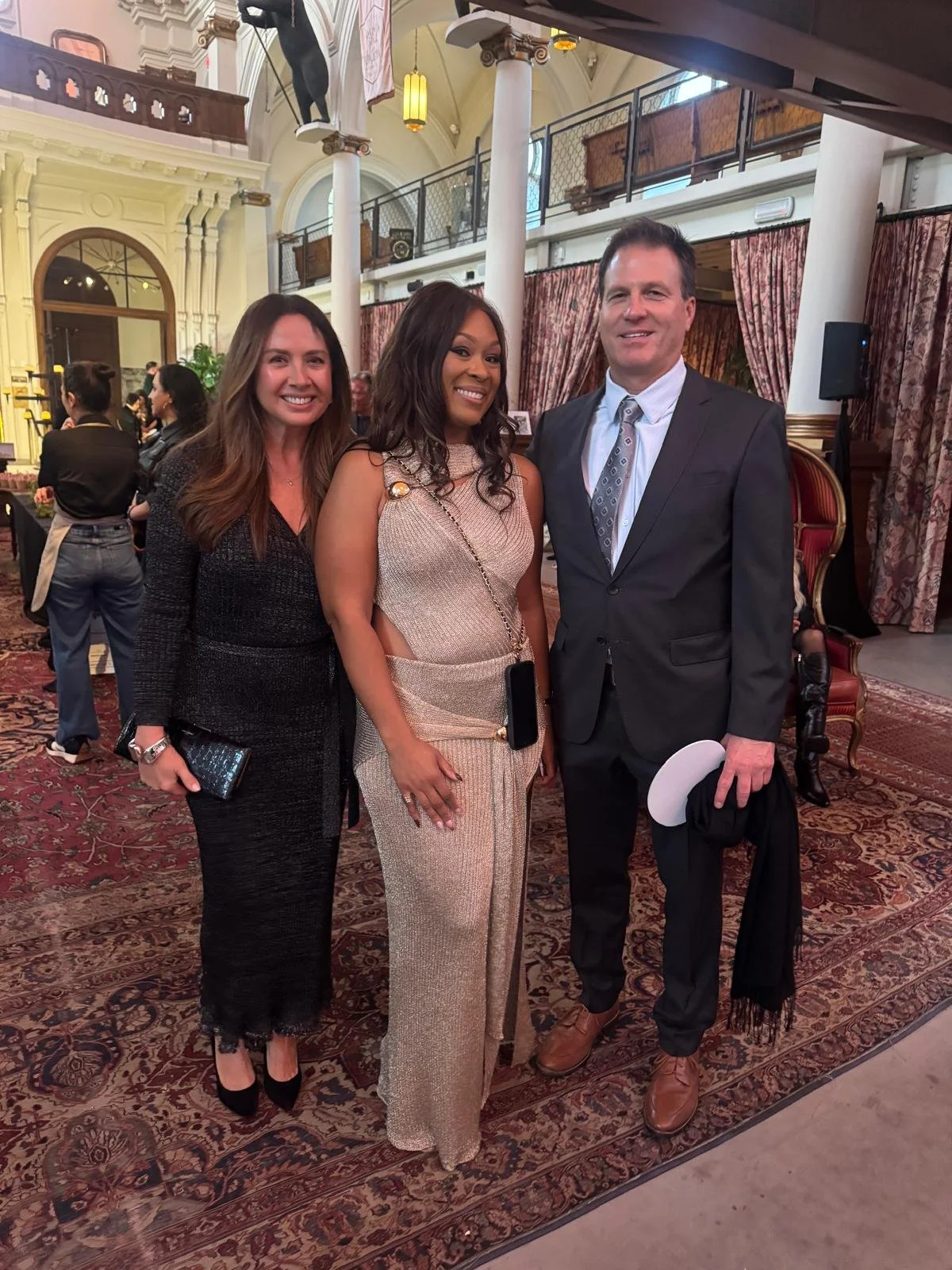 Three people dressed in formal attire standing inside a luxurious venue with ornate decor, drapes, and a patterned carpet.