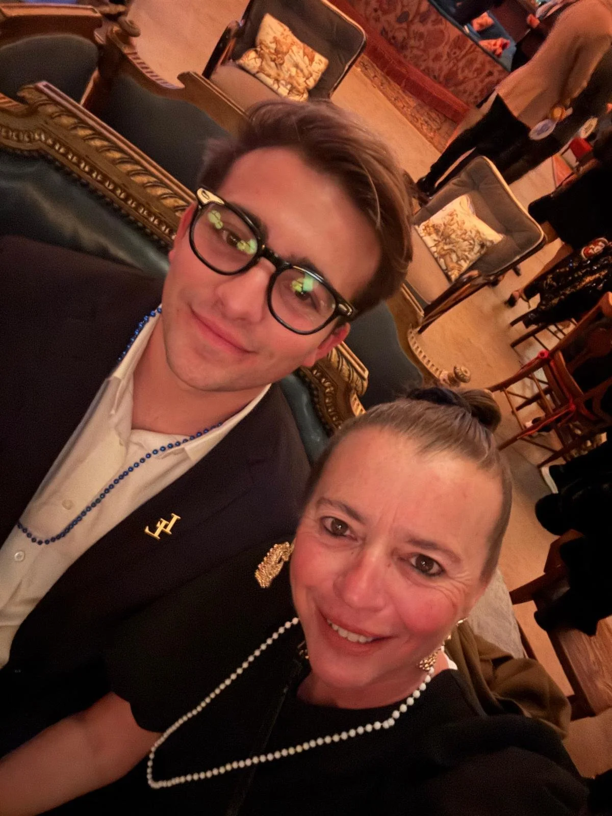 Two people taking a selfie in an elegant, vintage-style sitting area with chairs and floral cushions, with other guests in the background.