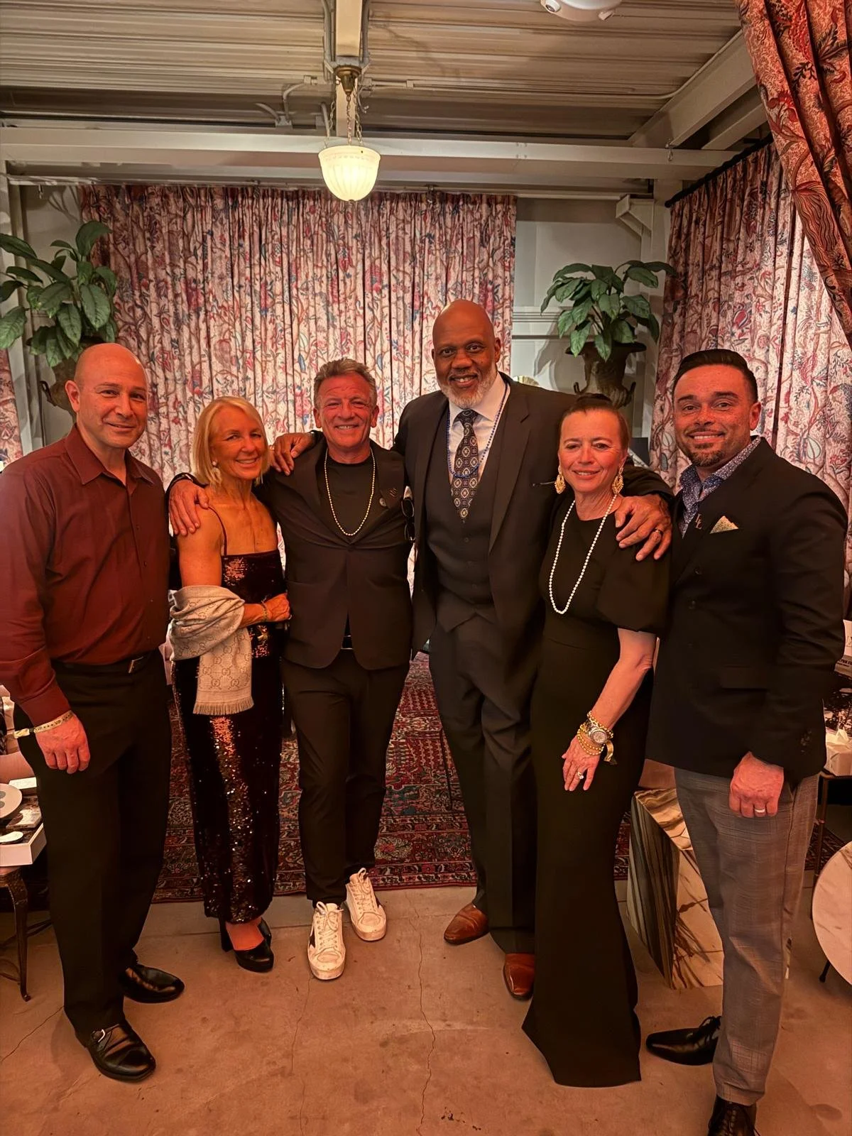 Group of six people dressed in formal attire, standing together in a room with floral curtains and indoor plants.