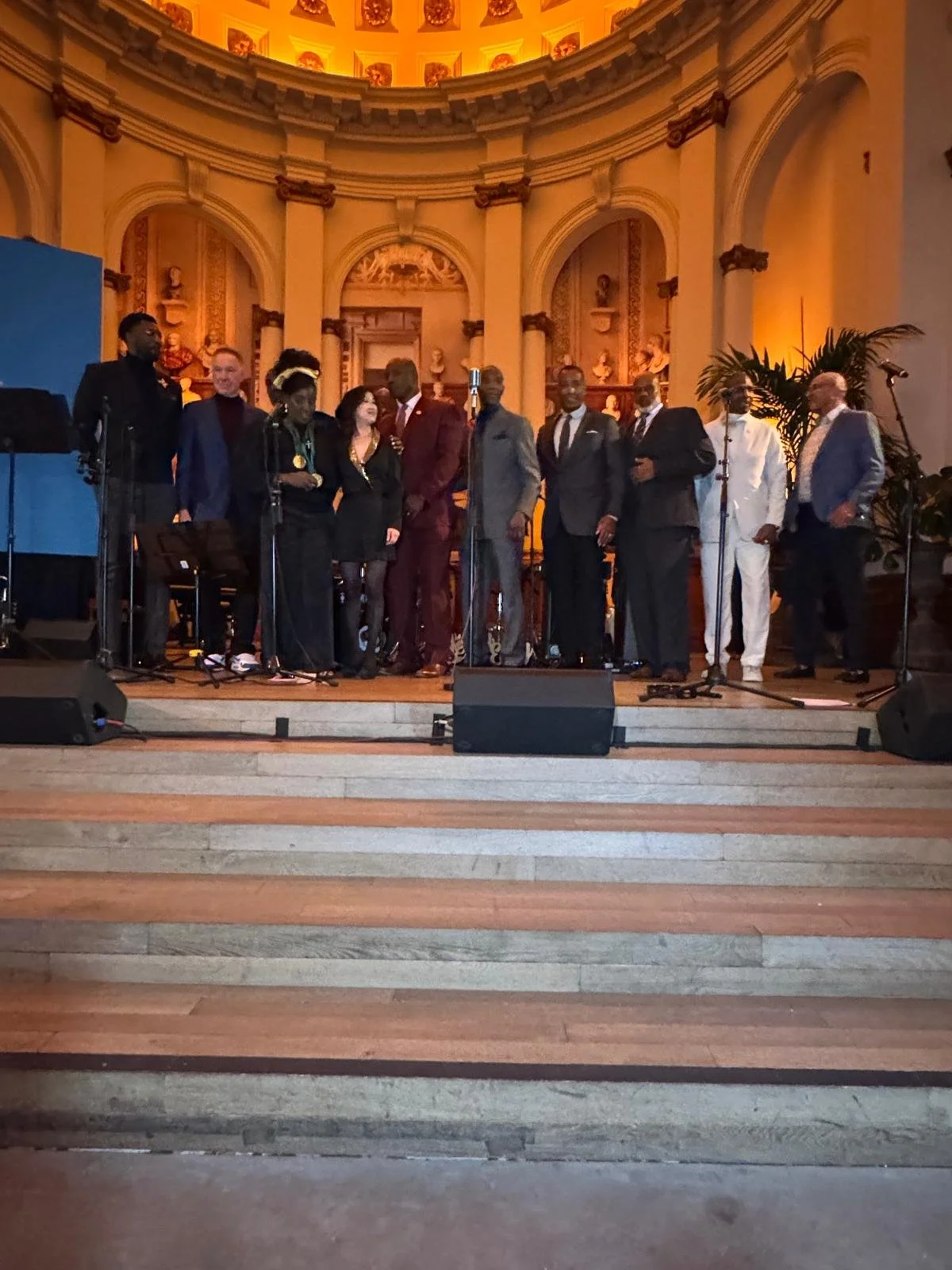 A group of ten people on stage in an elegant, ornately decorated hall with a domed ceiling and sculptures, participating in a formal event or performance.