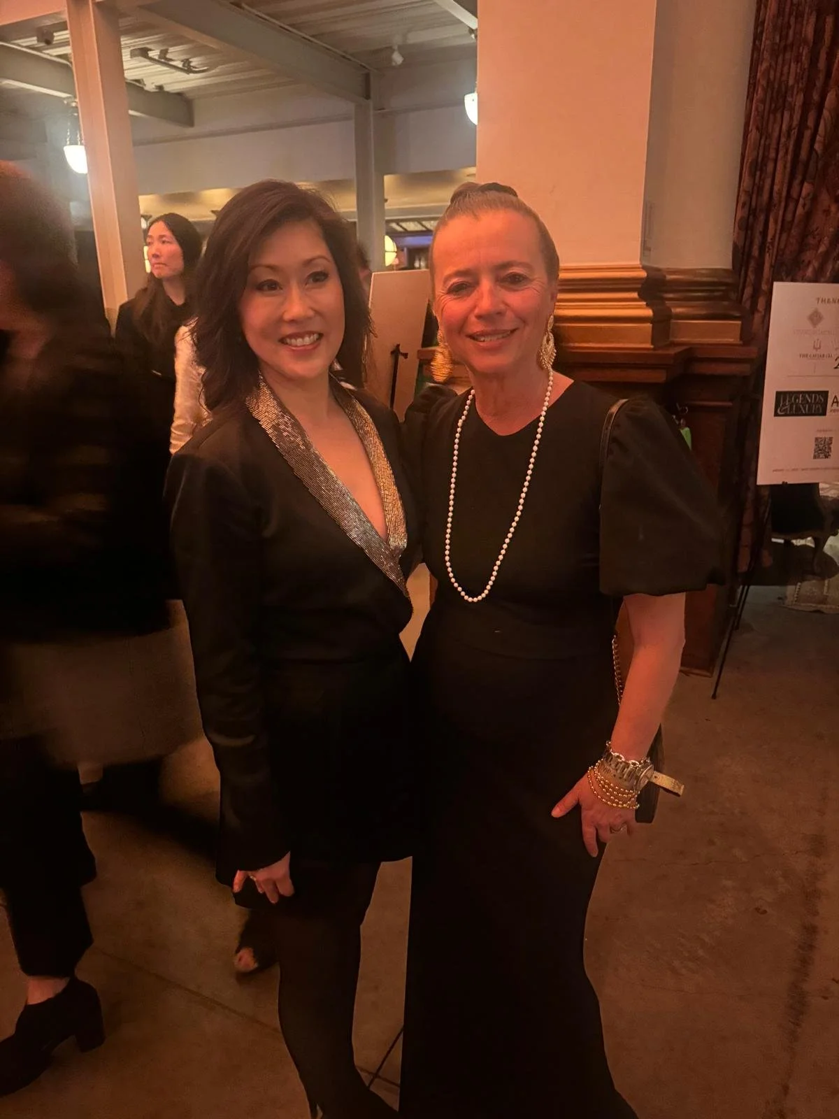 Two women in formal attire posing together at an indoor event, both smiling.