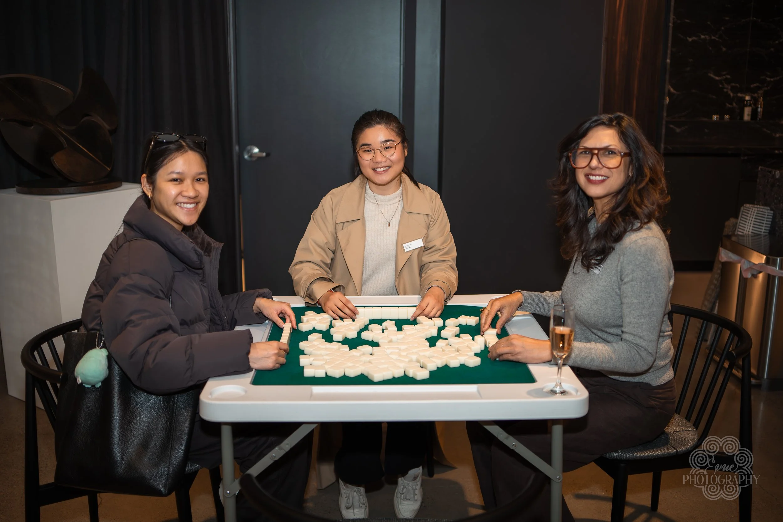 Three women playing mahjong at a table, smiling, with one holding a glass of champagne.