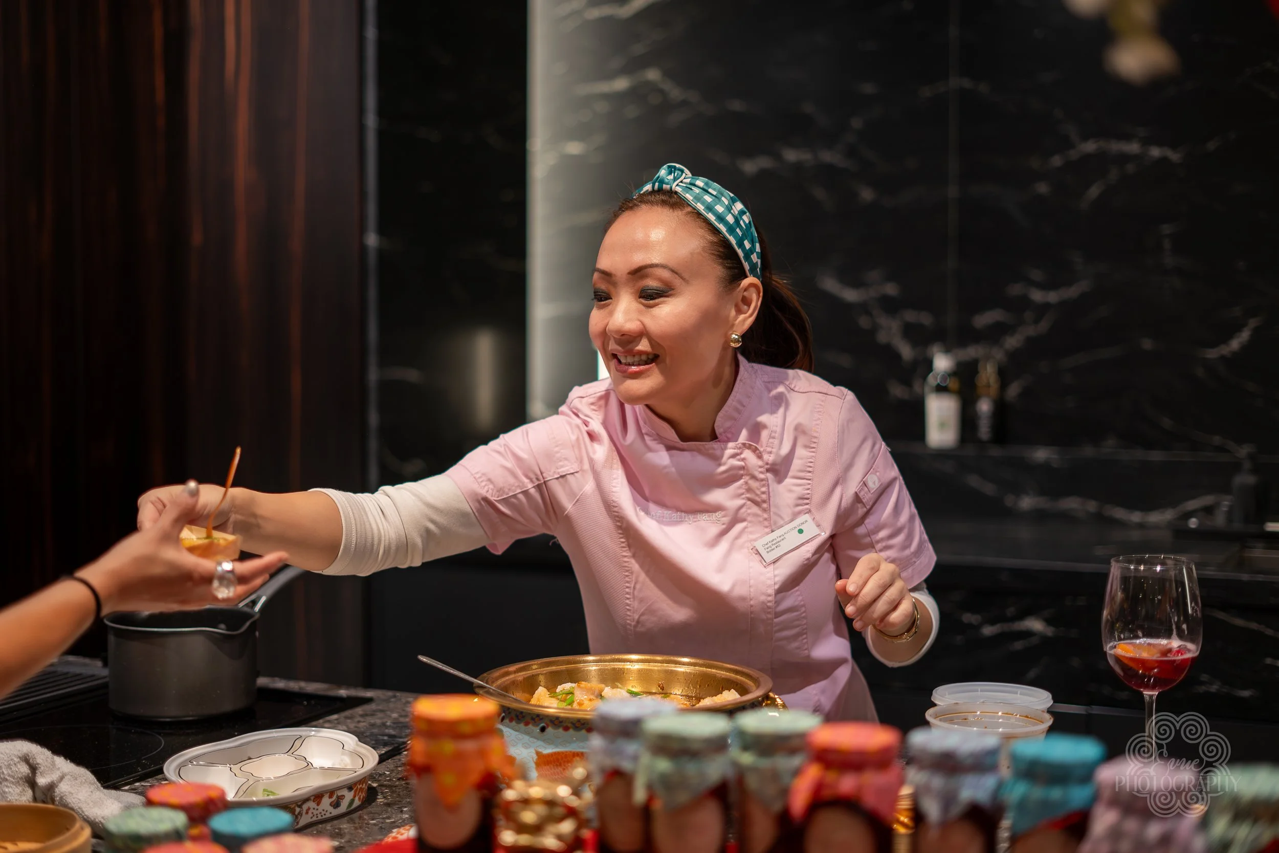 A woman wearing a pink chef's coat and a blue headband smiling as she hands a cupcake with orange frosting to someone. There are colorful baked goods and jars on the counter in front of her.