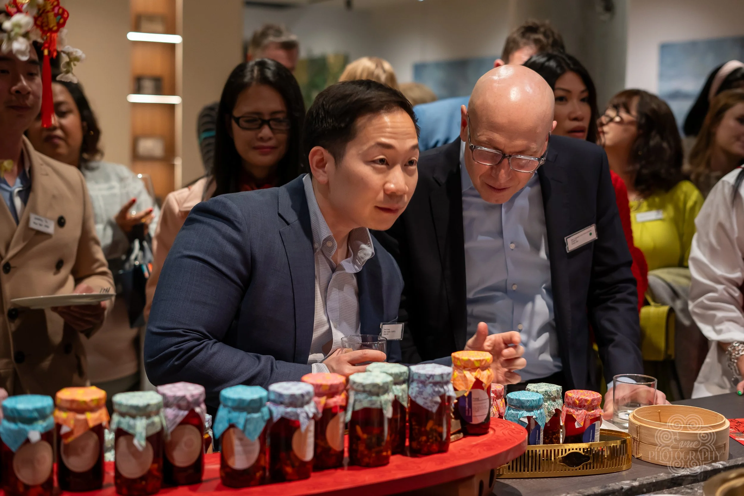 Two men in business attire examining jars of jam at a market or exhibition, with other people in the background.