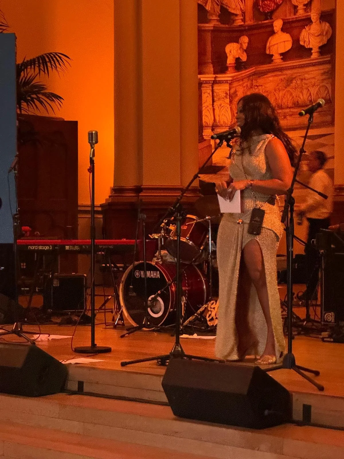 A woman standing on stage with a microphone, holding a piece of paper, surrounded by musical equipment including drums, a keyboard, and microphones, in a dimly lit venue with ornate decor.