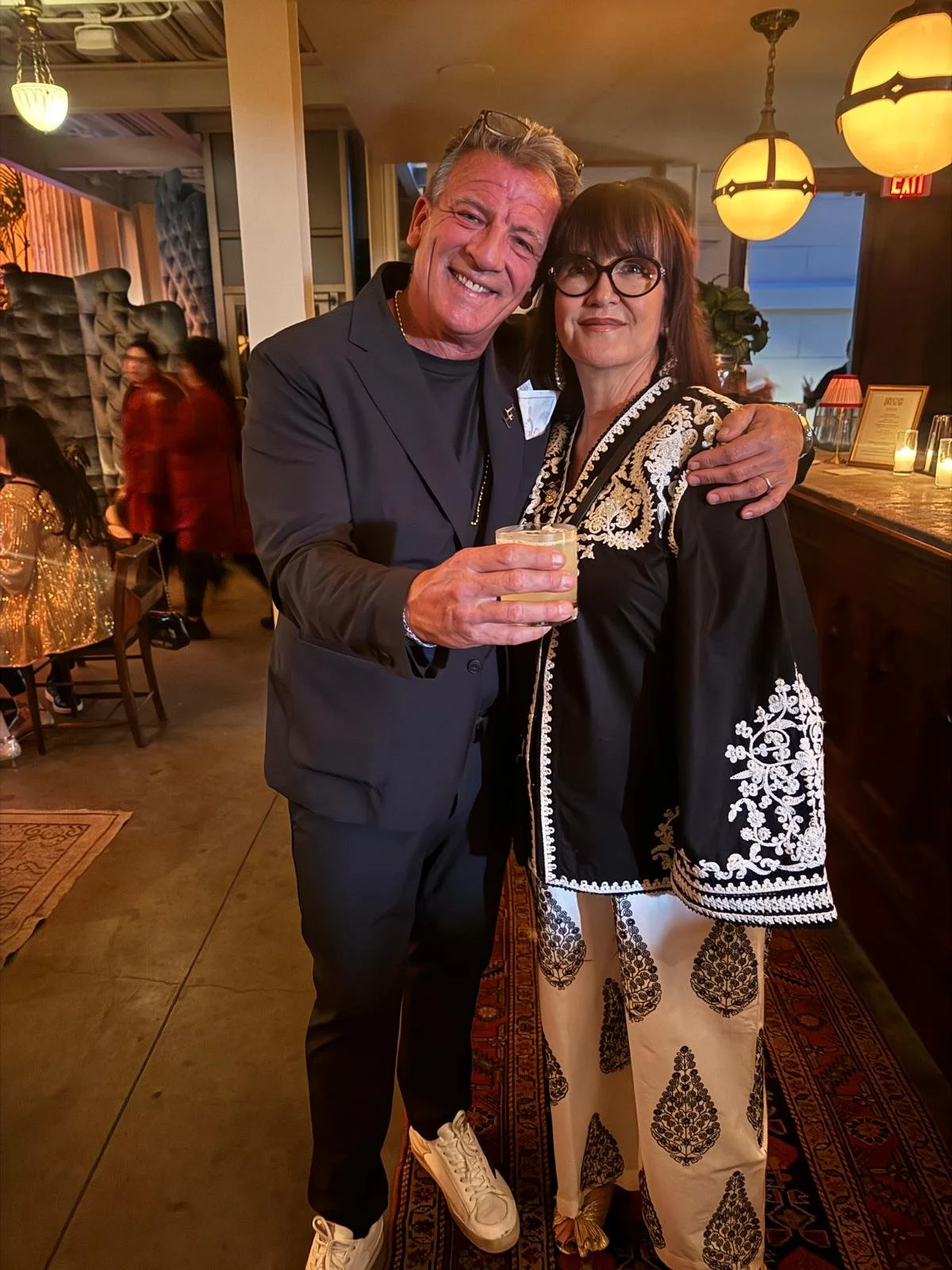 A man and woman smiling at a restaurant, holding a drink, with warm lighting and other patrons in the background.