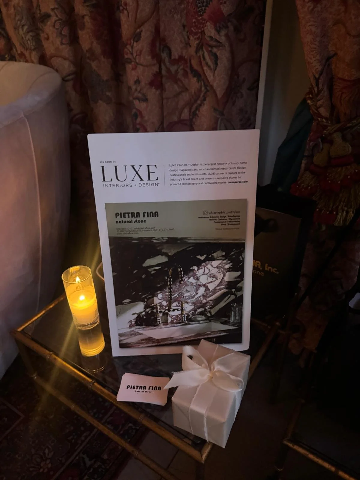 Display of interior design sample with a brochure, a candle, and a white gift box on a glass table in a cozy, ornately draped room.
