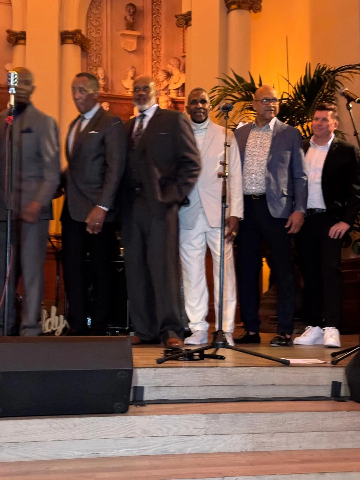 Six men dressed in formal suits standing on a stage with microphones and decorative background.