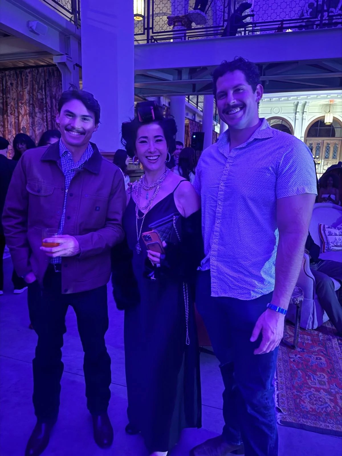 Three people smiling and posing together at a social event indoors, with a purple lighting ambiance and decorative interior architecture visible in the background.