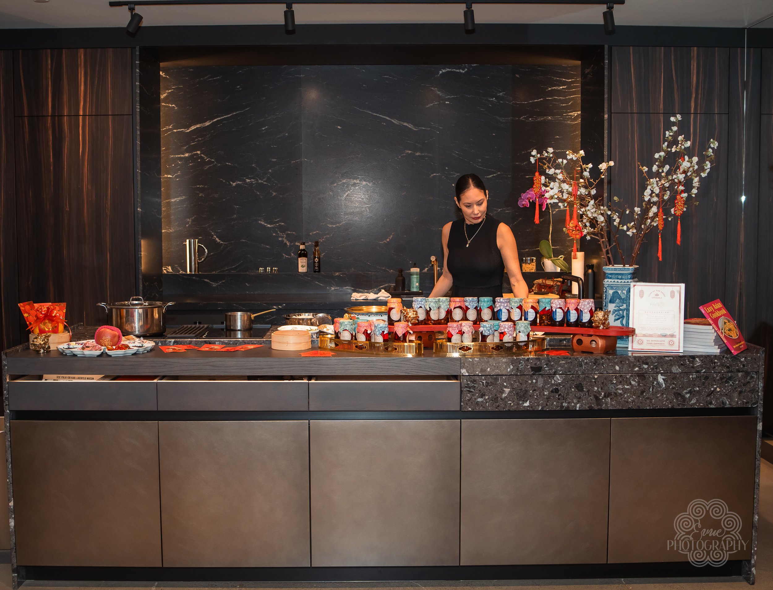 A woman standing behind a kitchen counter with various jars and decorative items, in a modern, dark-themed interior with black marble backsplash and wood-paneled walls.