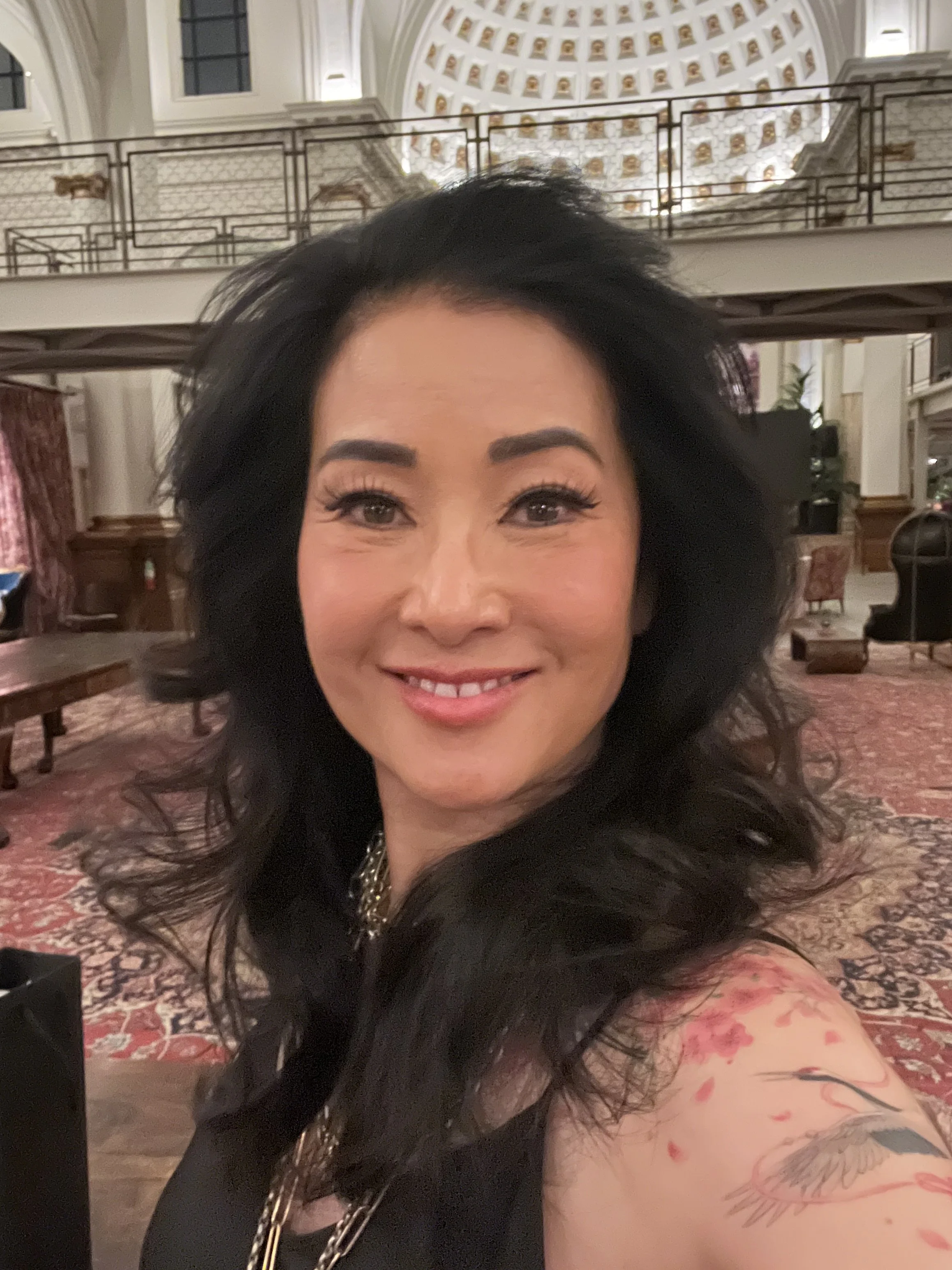 A woman with long dark hair and tattoos taking a selfie inside a grand, ornate building with detailed architectural features.
