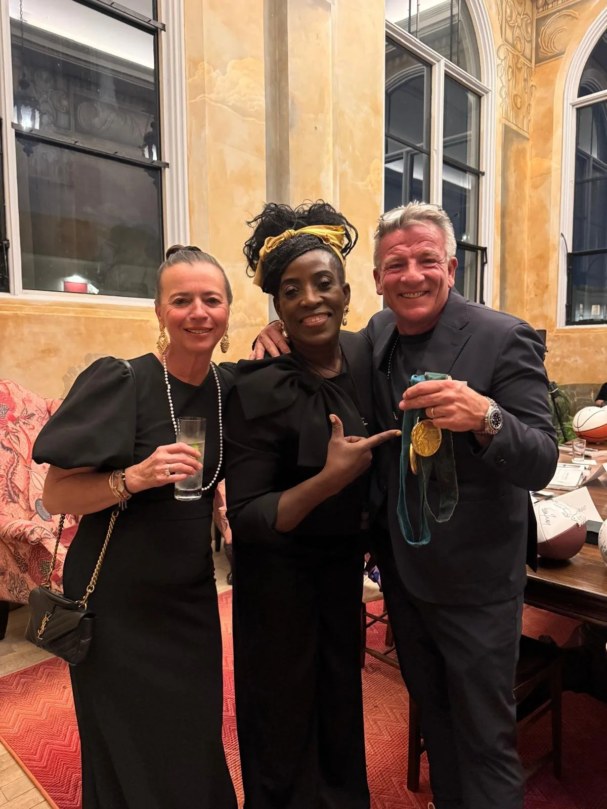 Three people smiling and posing for a photo at a celebration or formal event. The woman on the left is holding a drink, the woman in the middle is pointing at the man on the right who is holding medals.