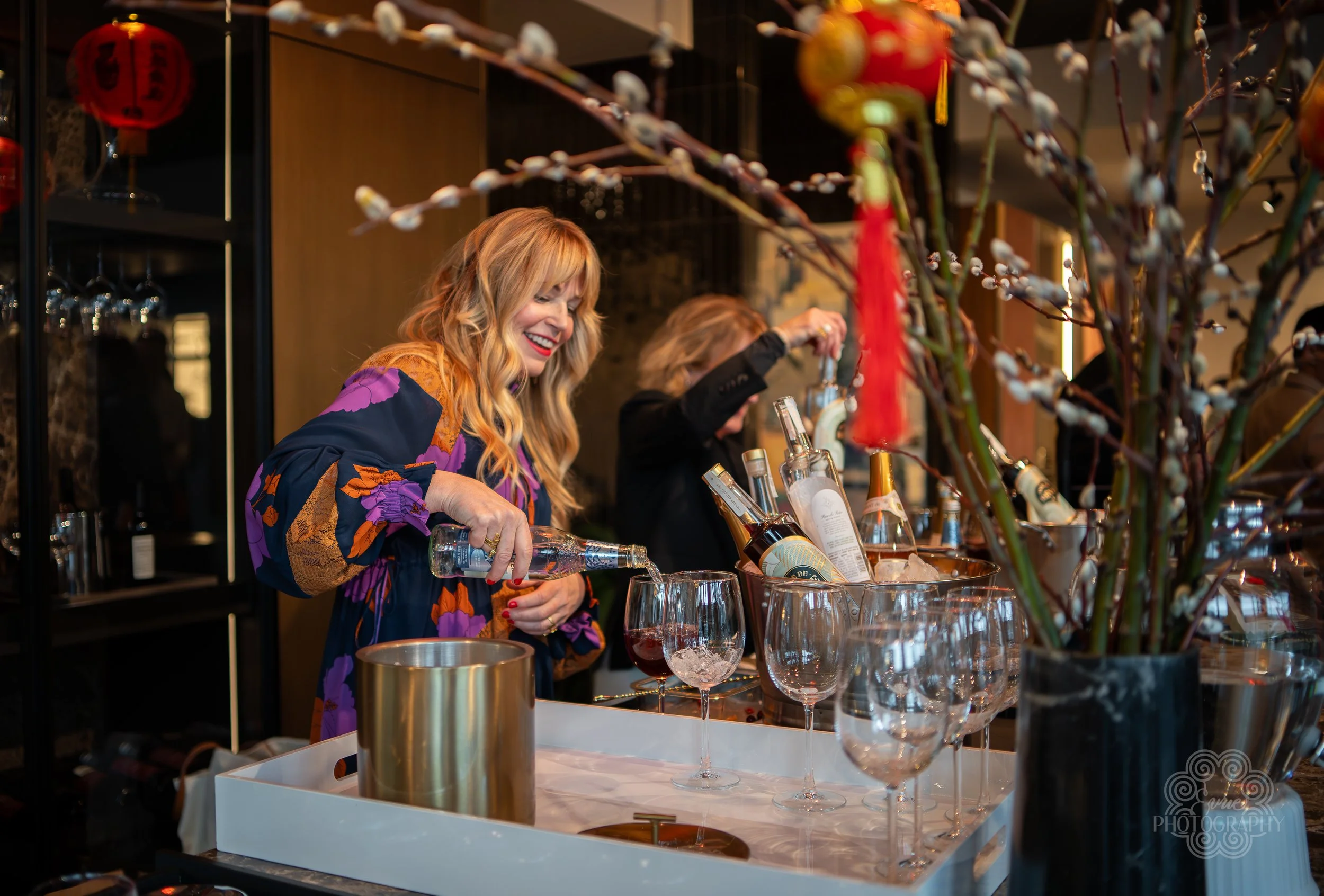 Women preparing drinks at a bar during a festive celebration with decorative branches and lanterns.