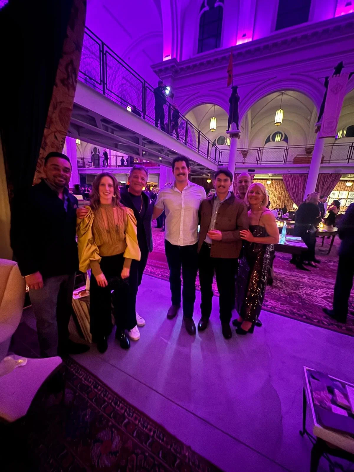 Group of seven people posing together at an event in an elegant venue with purple lighting, high ceilings, and decorative architecture.