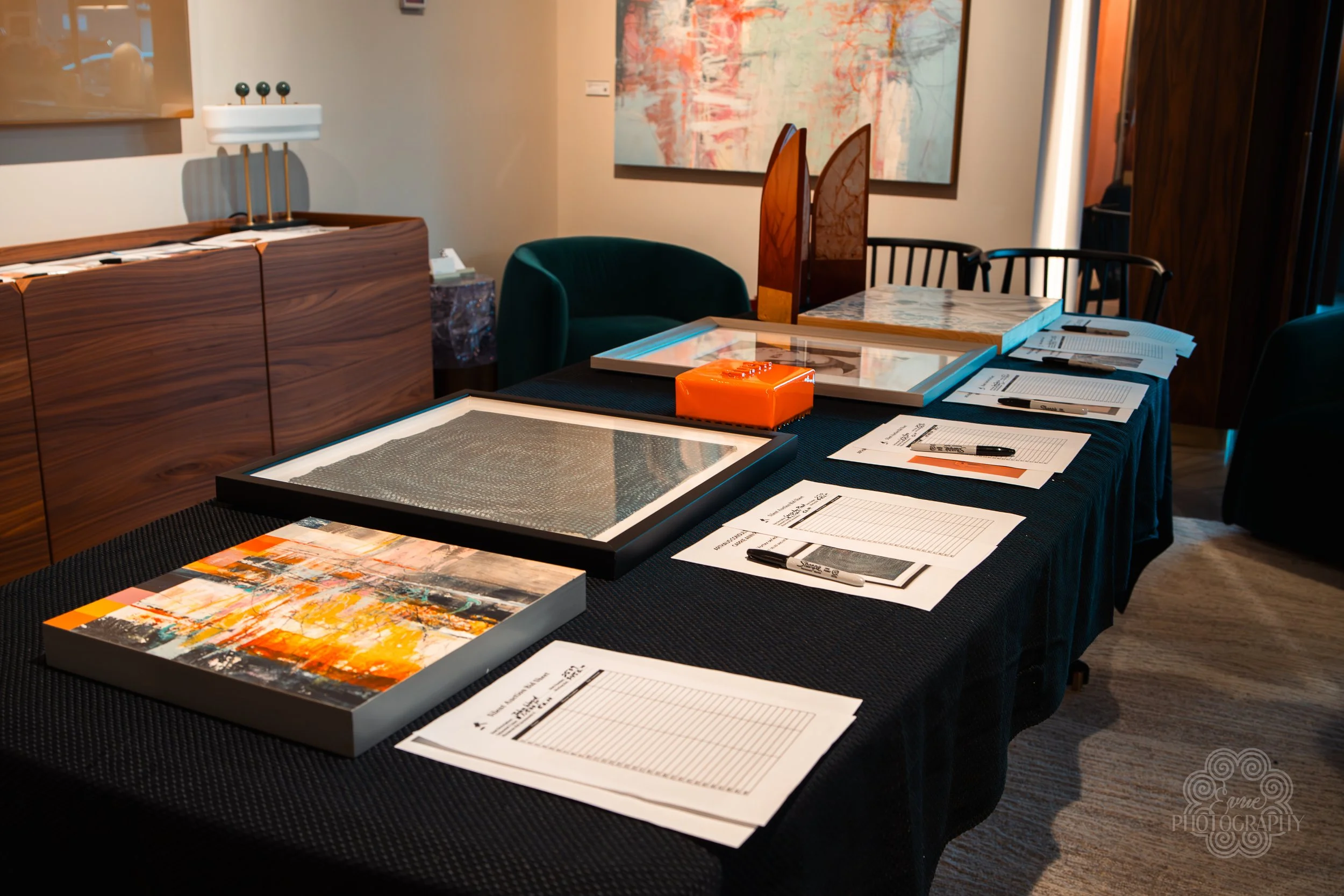 A conference or meeting table set up with framed artwork, papers, and pens, in a room with decorative chairs, wooden furniture, and abstract artwork on the wall.