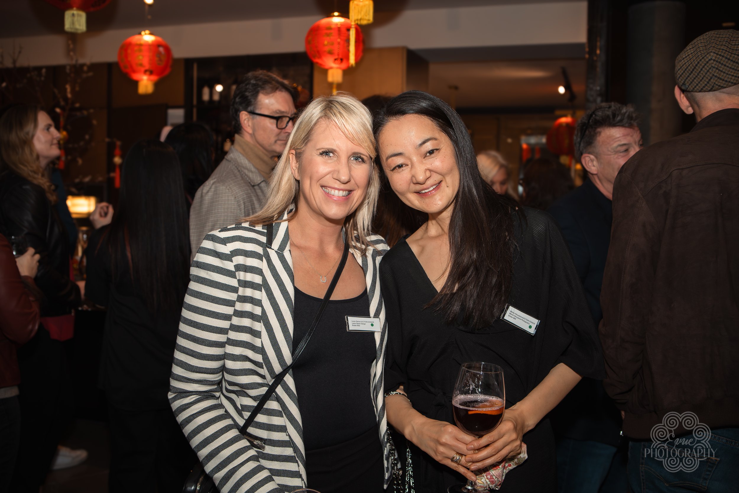 Two women smiling at a social event, one holding a glass of red wine, with Chinese lantern decorations hanging above in the background.