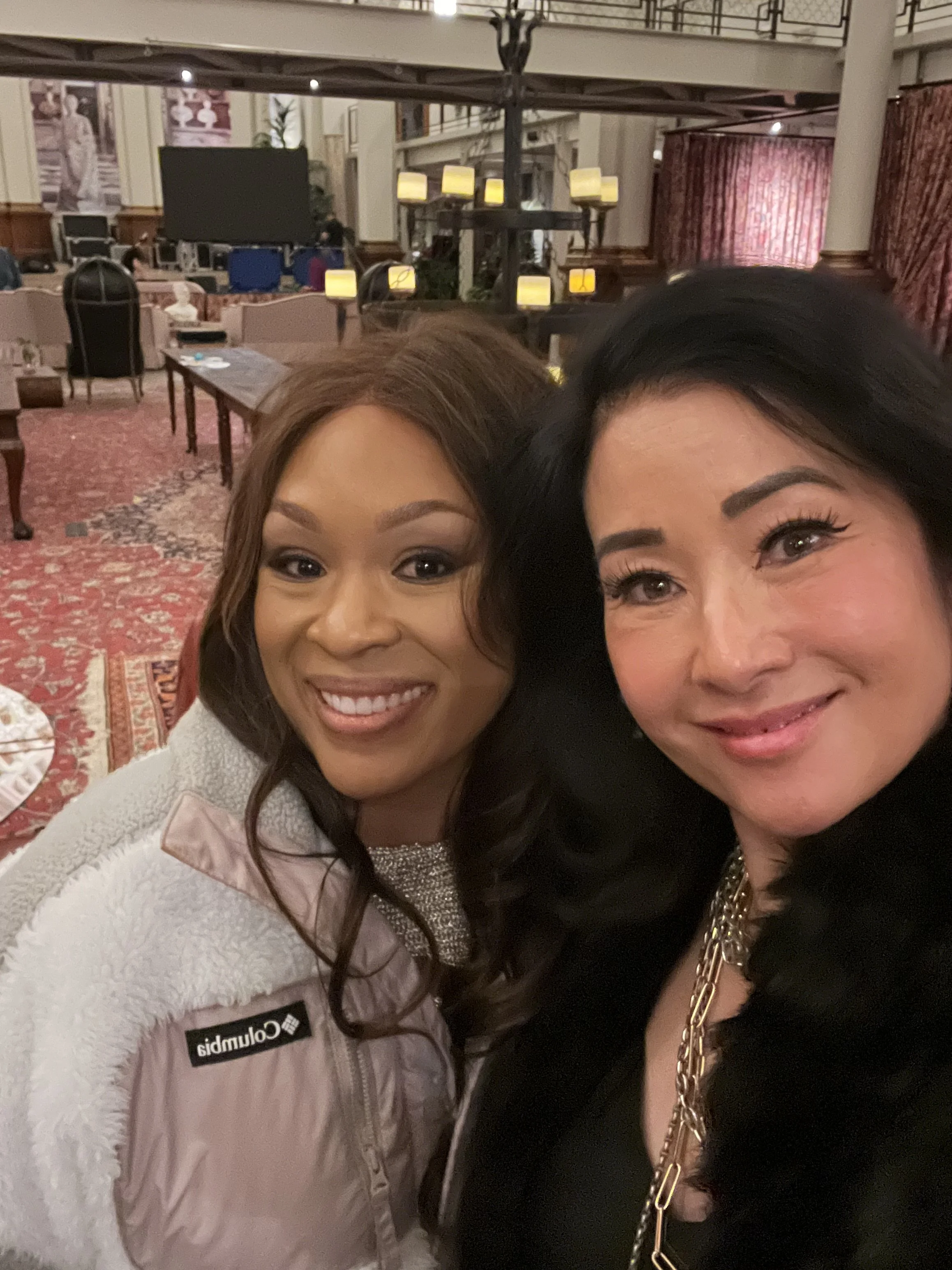 Two women smiling for a selfie in an elegant, warmly lit indoor setting with patterned carpet, tables, chairs, and decorative hanging lights.