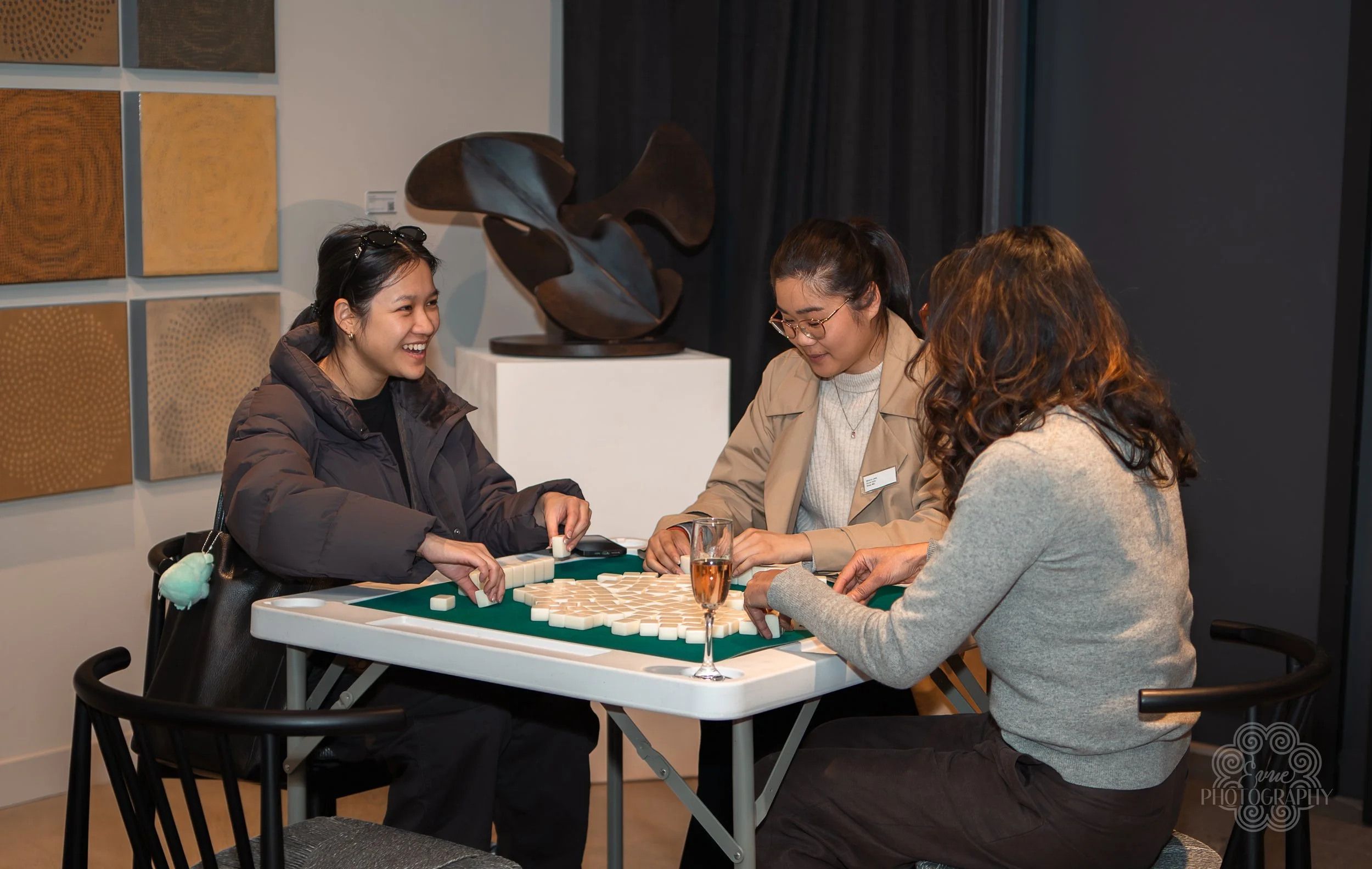 Three women playing a game of dominoes at a table, smiling and enjoying each other's company. One woman has curly hair, another is wearing glasses and a beige coat, and the third woman has sunglasses on her head and a gray coat. There is a glass of r