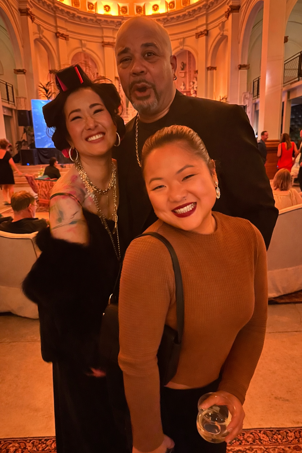 Three people smiling at a formal event or party in an elegant, warmly lit hall with ornate decoration and other guests seated in the background.