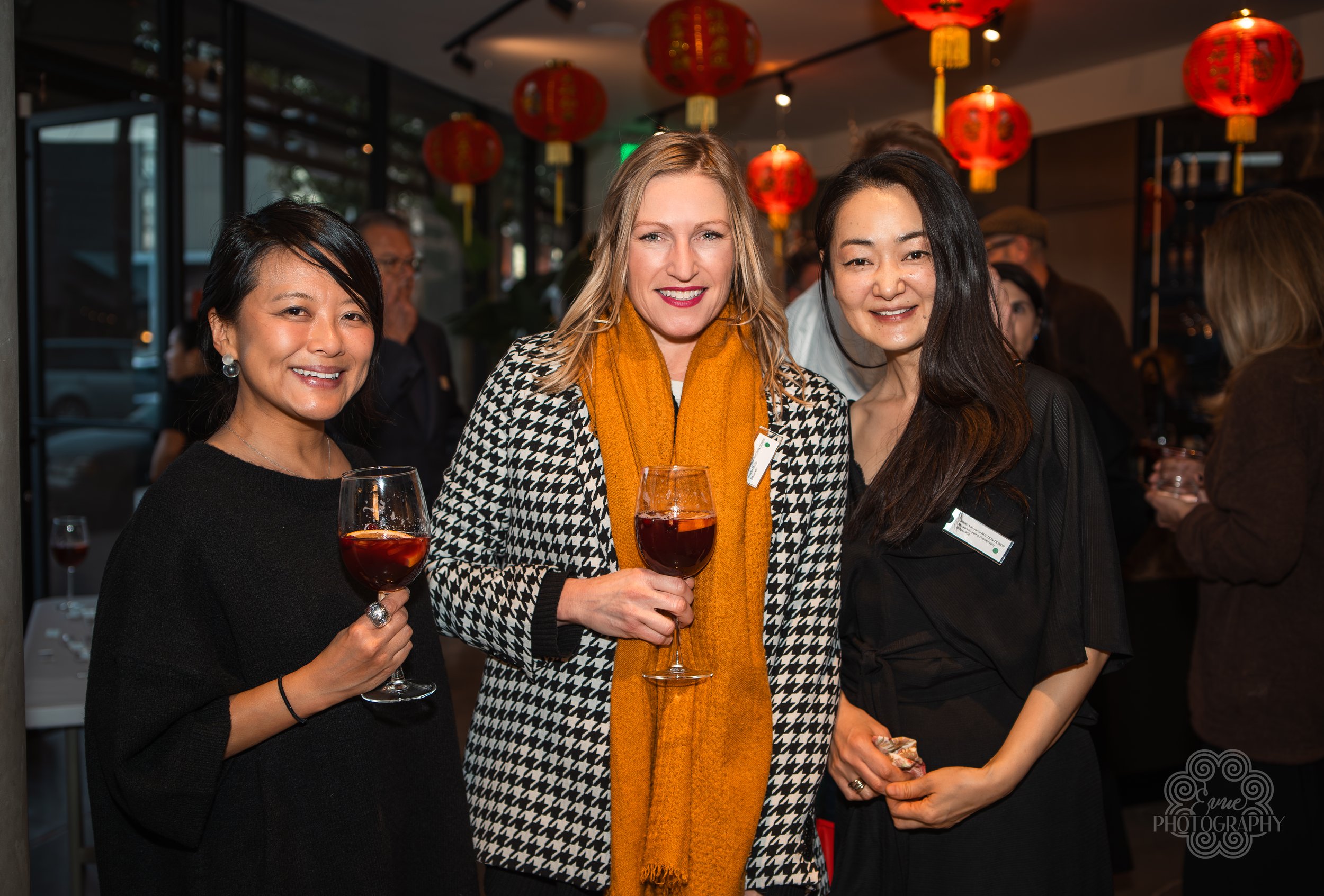Three women at a social gathering, holding glasses of red wine, smiling at the camera, with red lantern decorations in the background.