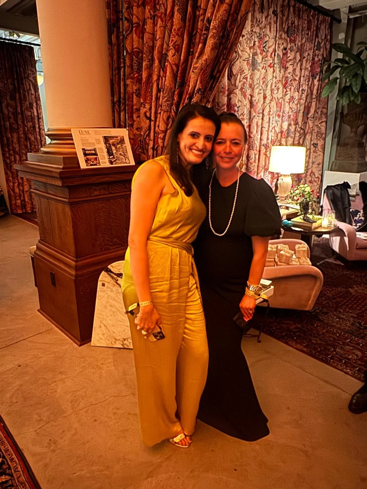 Two women standing close together, smiling, in a warmly lit room with floral curtains and furniture in the background.