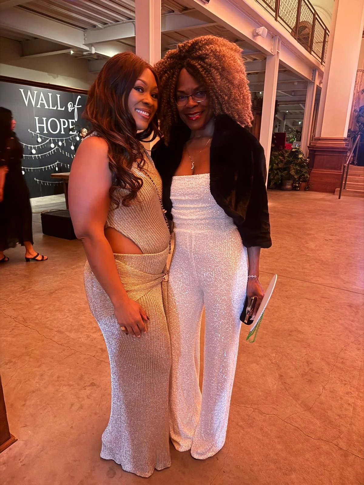 Two women dressed in shiny elegant outfits pose together inside a venue with a sign that reads 'Wall of Hope' in the background.