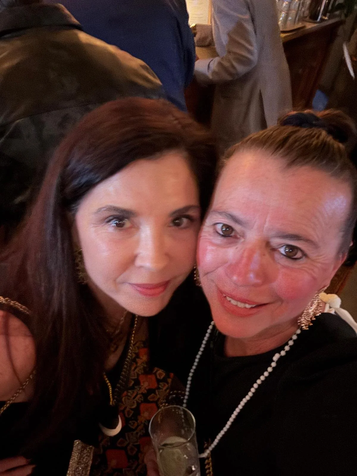 Two women taking a selfie at a social event, smiling, holding a glass of wine, with a cozy indoor setting in the background.