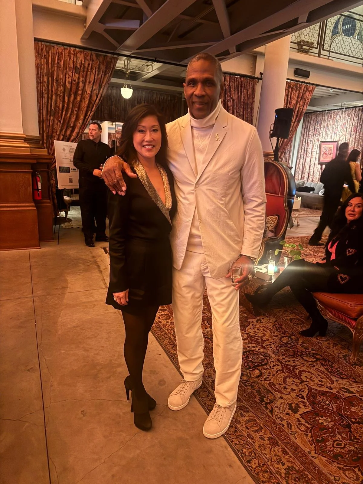 A woman in a black dress and black heels standing next to a tall man in a white suit, both smiling at the camera in an elegant indoor setting with floral curtains, a patterned rug, and dim lighting.