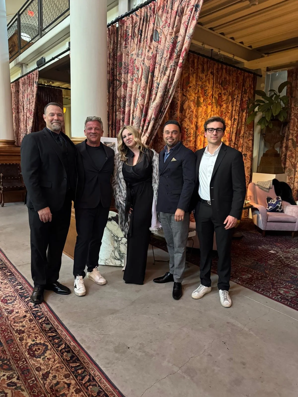 Group of five people dressed in formal attire, standing in an elegant interior with floral curtains, a plant, a marble-topped table, and vintage furniture.
