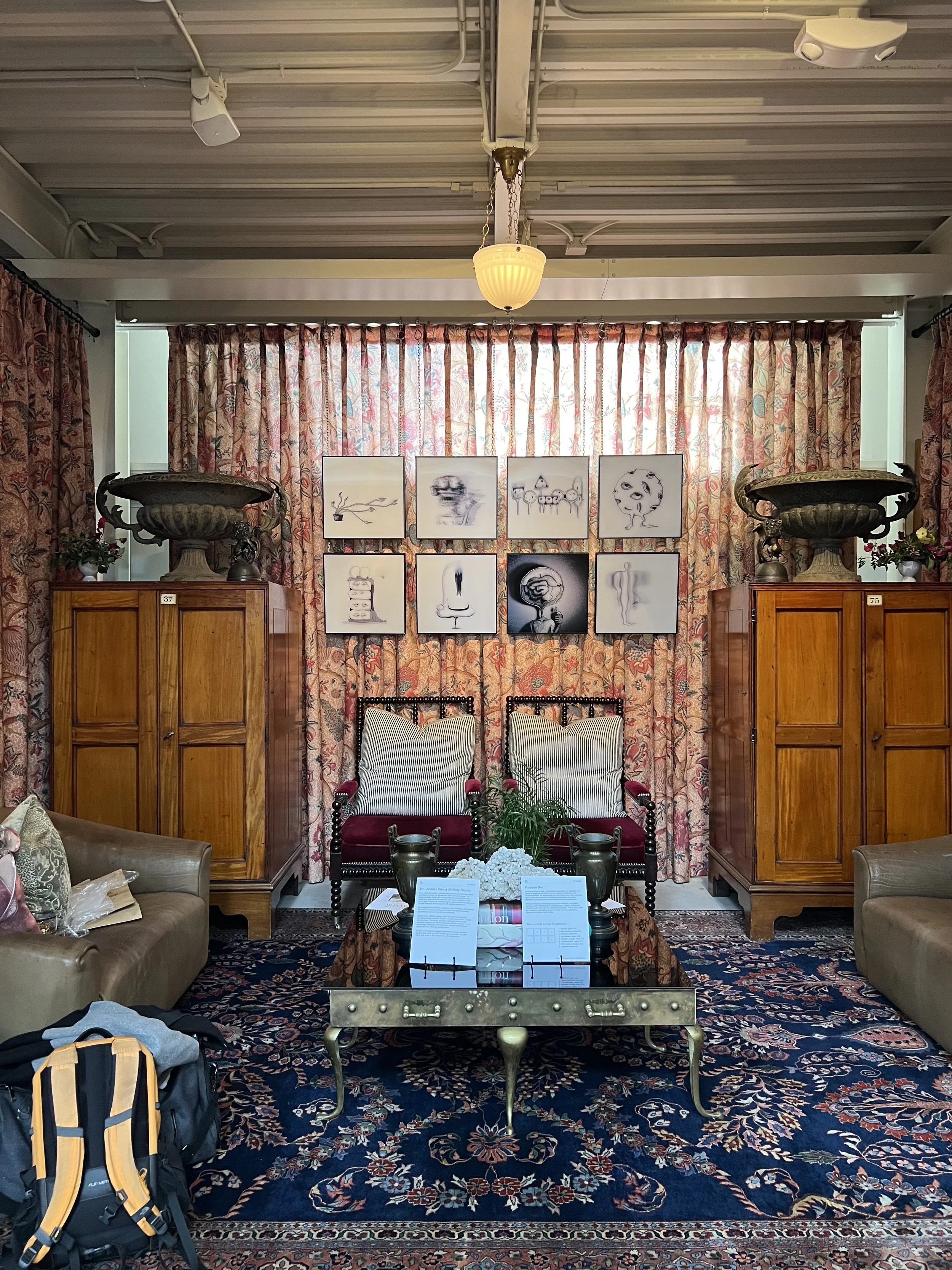 A cozy living room with two wooden cabinets, a floral curtain backdrop, two armchairs, a gold-colored coffee table with informational signs, and framed black-and-white artistic photos on the curtain. There is a backpack near the sofa on the left.