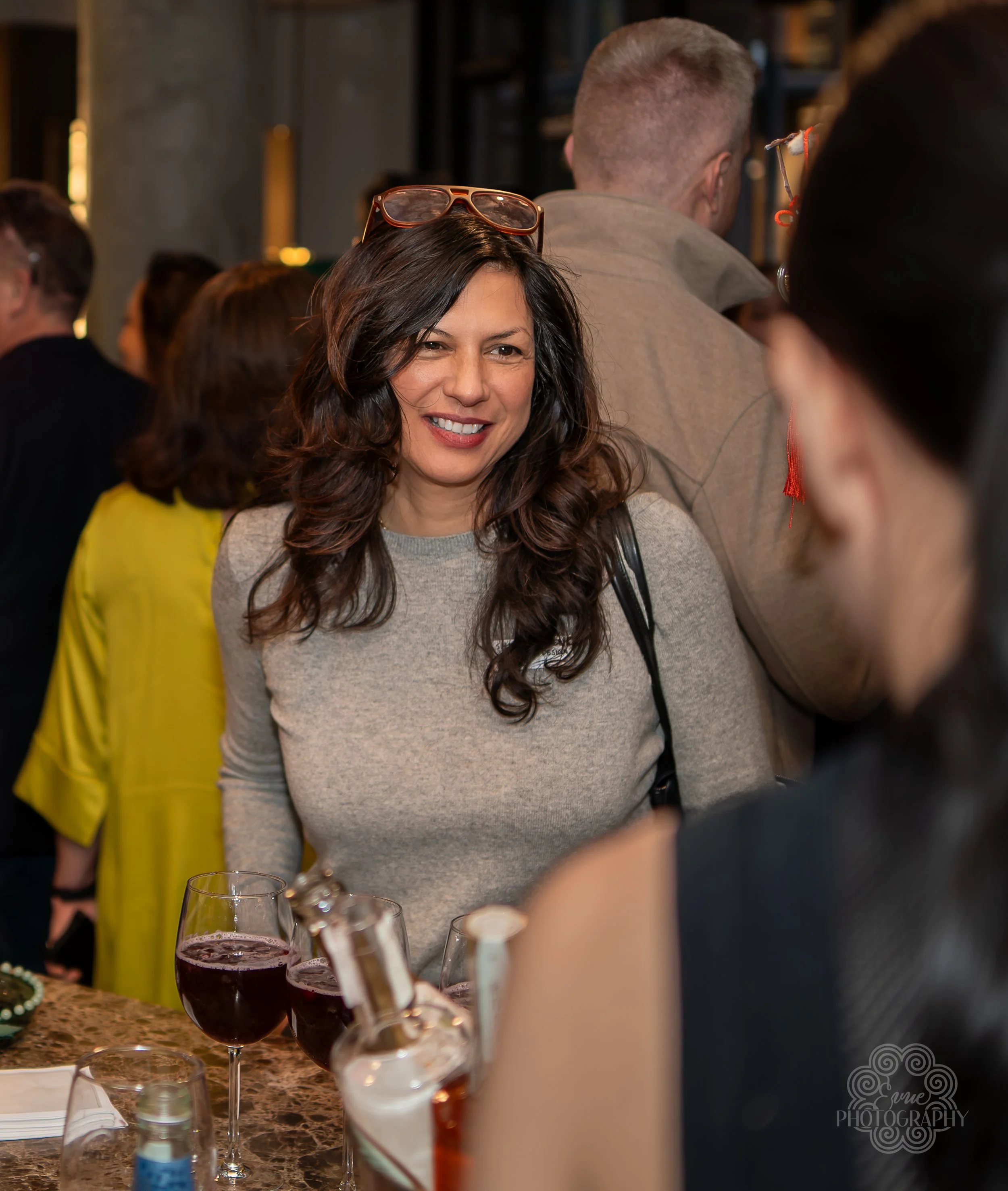 A woman with long curly brown hair smiling in a social gathering, standing at a table with glasses of red wine, in a crowded indoor setting.