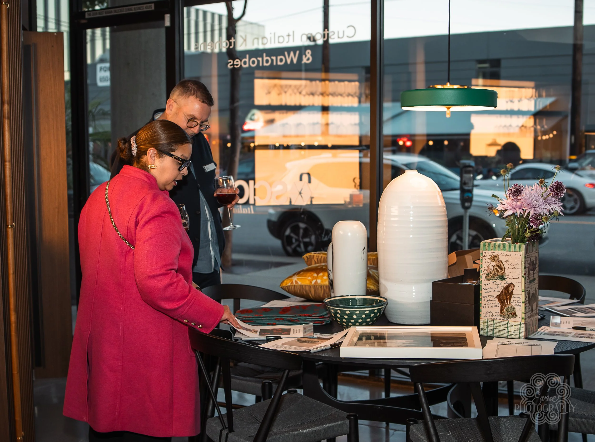 Two people in a store looking at home decor items on a black table, with large vases, a bowl, and a picture frame, inside a space with large window showing cars outside.